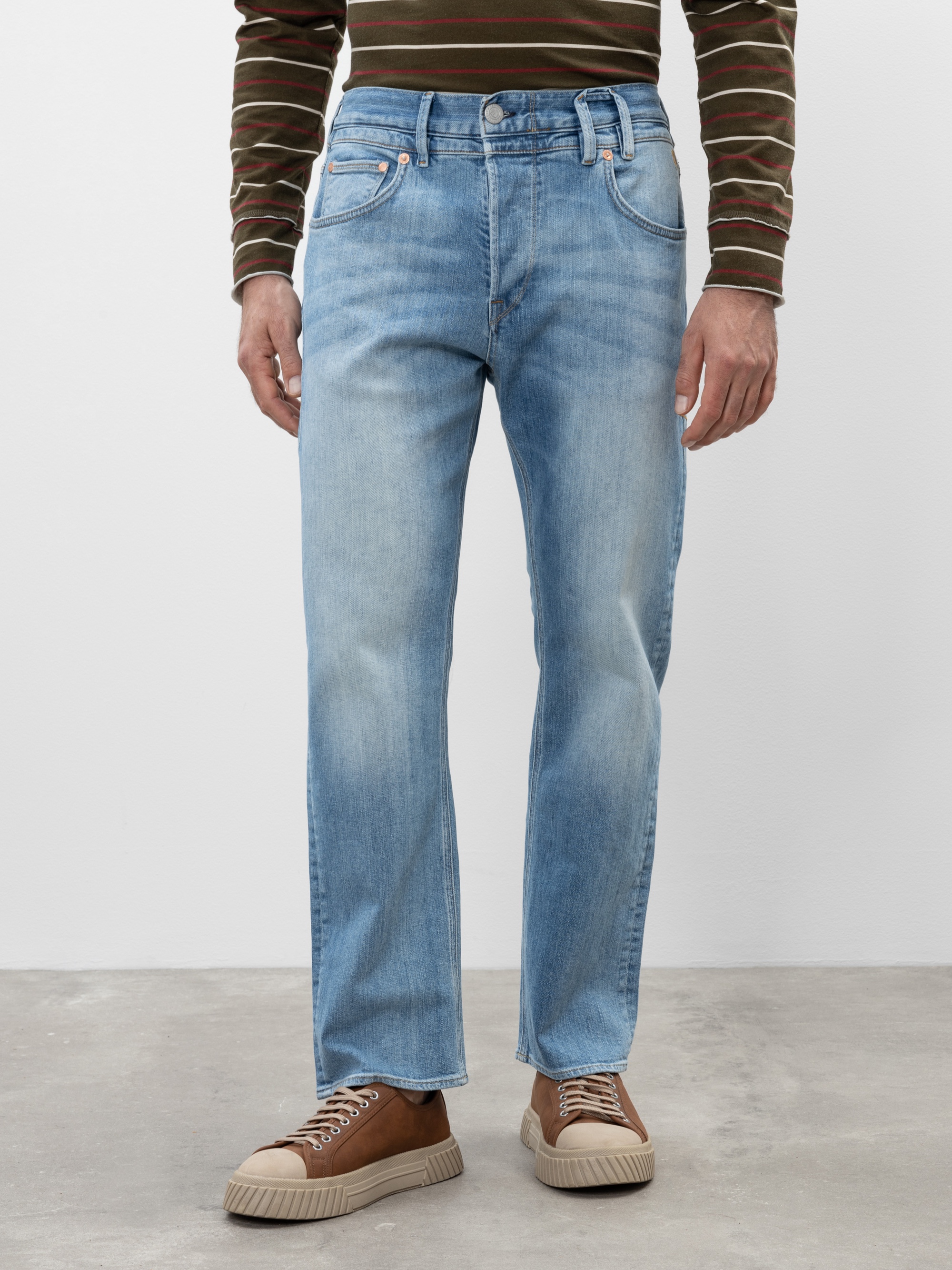 Eine Person, die hellblaue Jeans mit geradem Bein, ein braun-grün gestreiftes langärmeliges Hemd und hellbraune Turnschuhe trägt, steht auf einem Betonboden vor einem schlichten weißen Hintergrund.