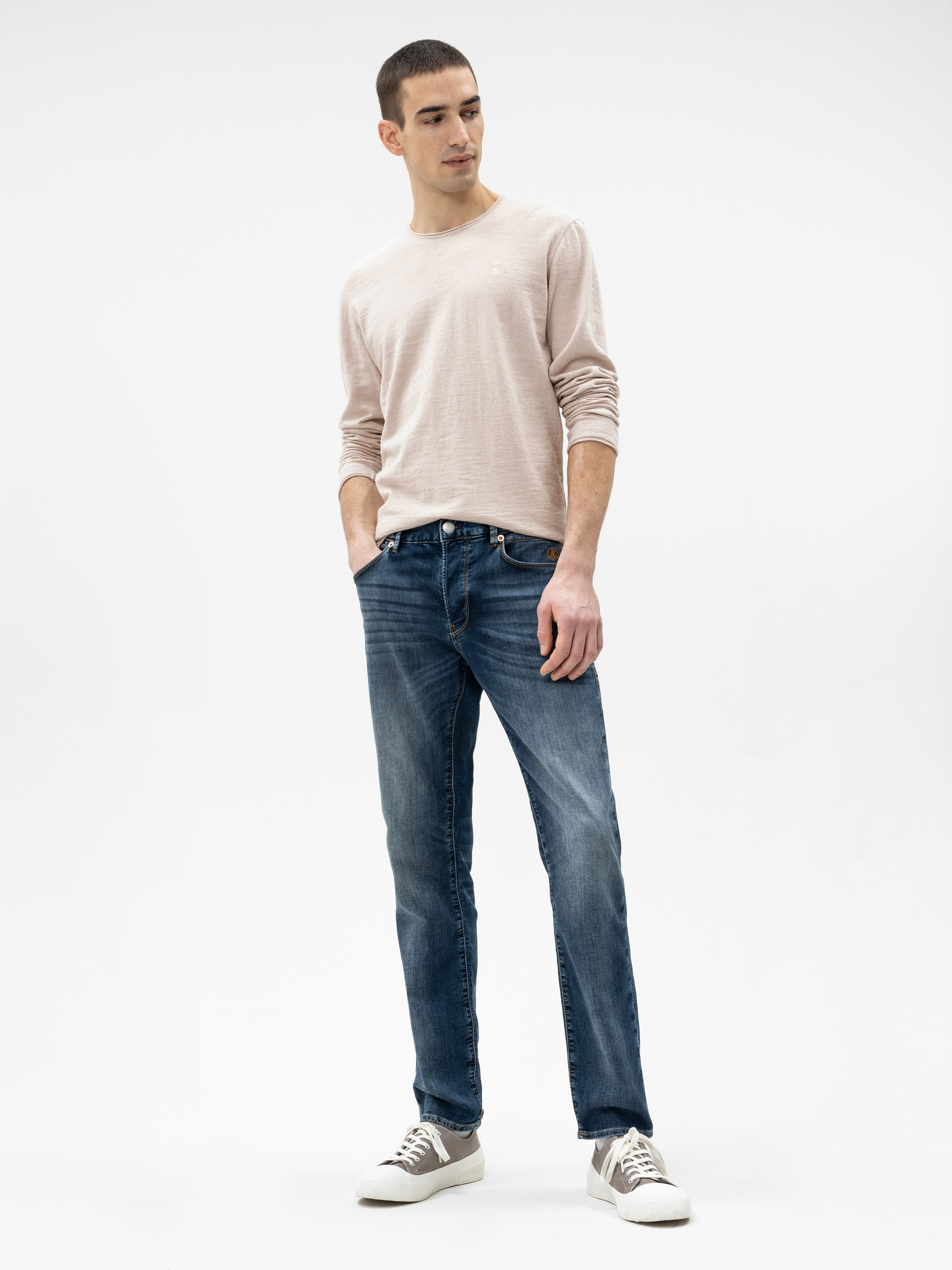 Ein Mann steht vor einem schlichten weißen Hintergrund und trägt ein hellbeiges langärmeliges Hemd, blaue Jeans und graue Turnschuhe. Er hat kurzes dunkles Haar, die Hände in den Taschen und schaut leicht zur Seite.
