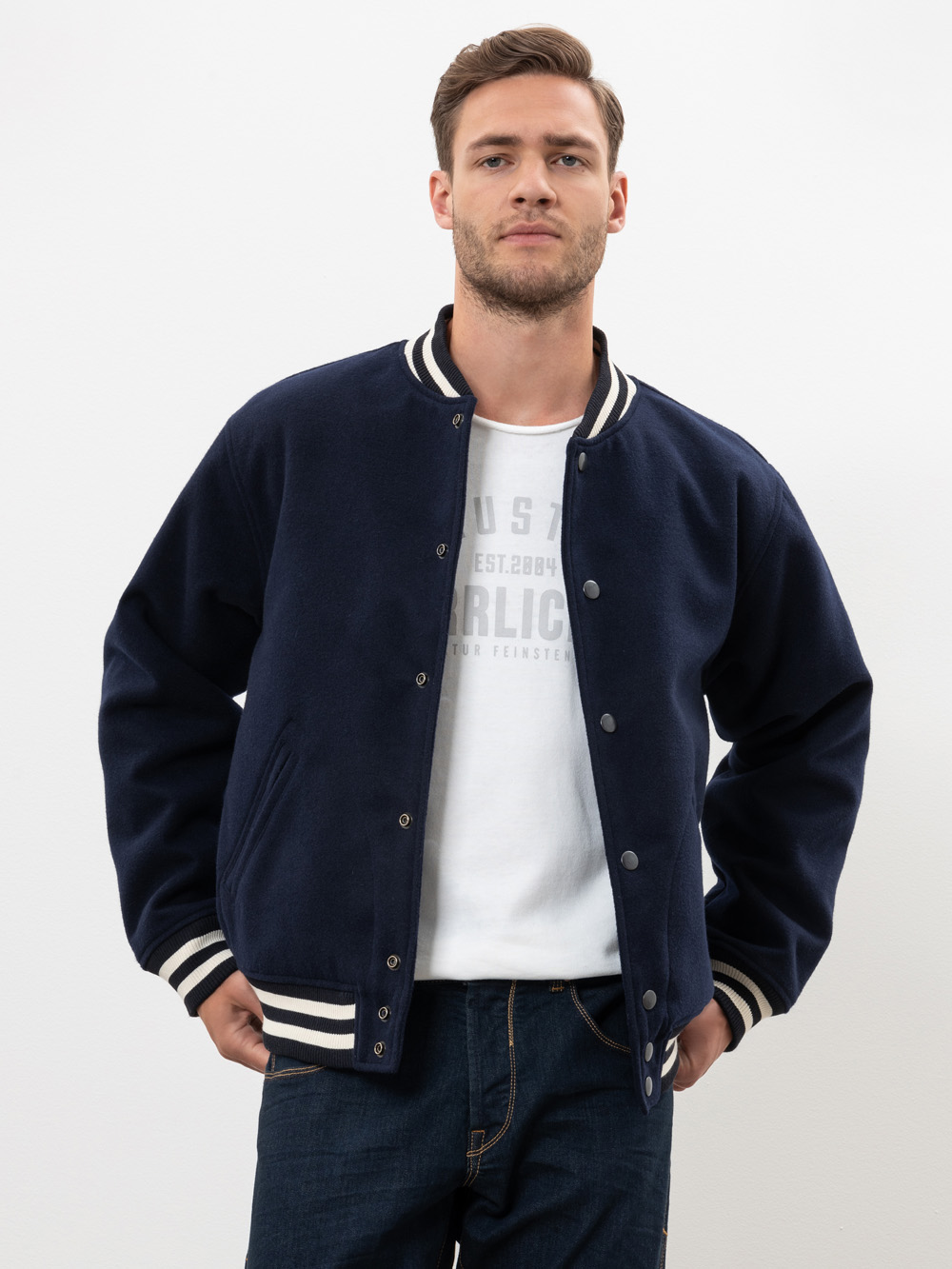 Ein Mann in einer marineblauen Sportjacke mit weiß gestreiften Manschetten und Kragen steht vor einem schlichten weißen Hintergrund. Er hat kurze Haare, einen gestutzten Bart und trägt ein weißes T-Shirt und blaue Jeans.