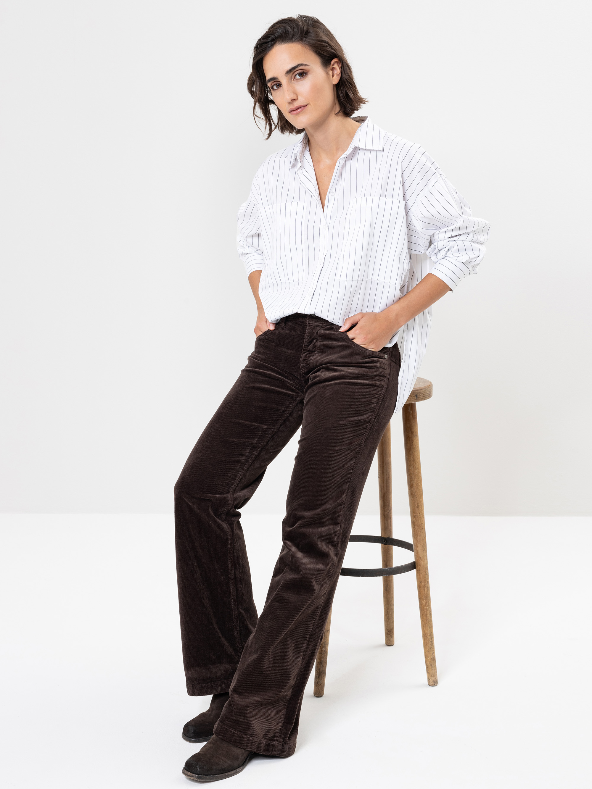 Eine Person mit kurzen braunen Haaren trägt ein weiß gestreiftes Button-up-Hemd und eine dunkelbraune Samthose und sitzt lässig auf einem Holzhocker vor einem schlichten weißen Hintergrund.