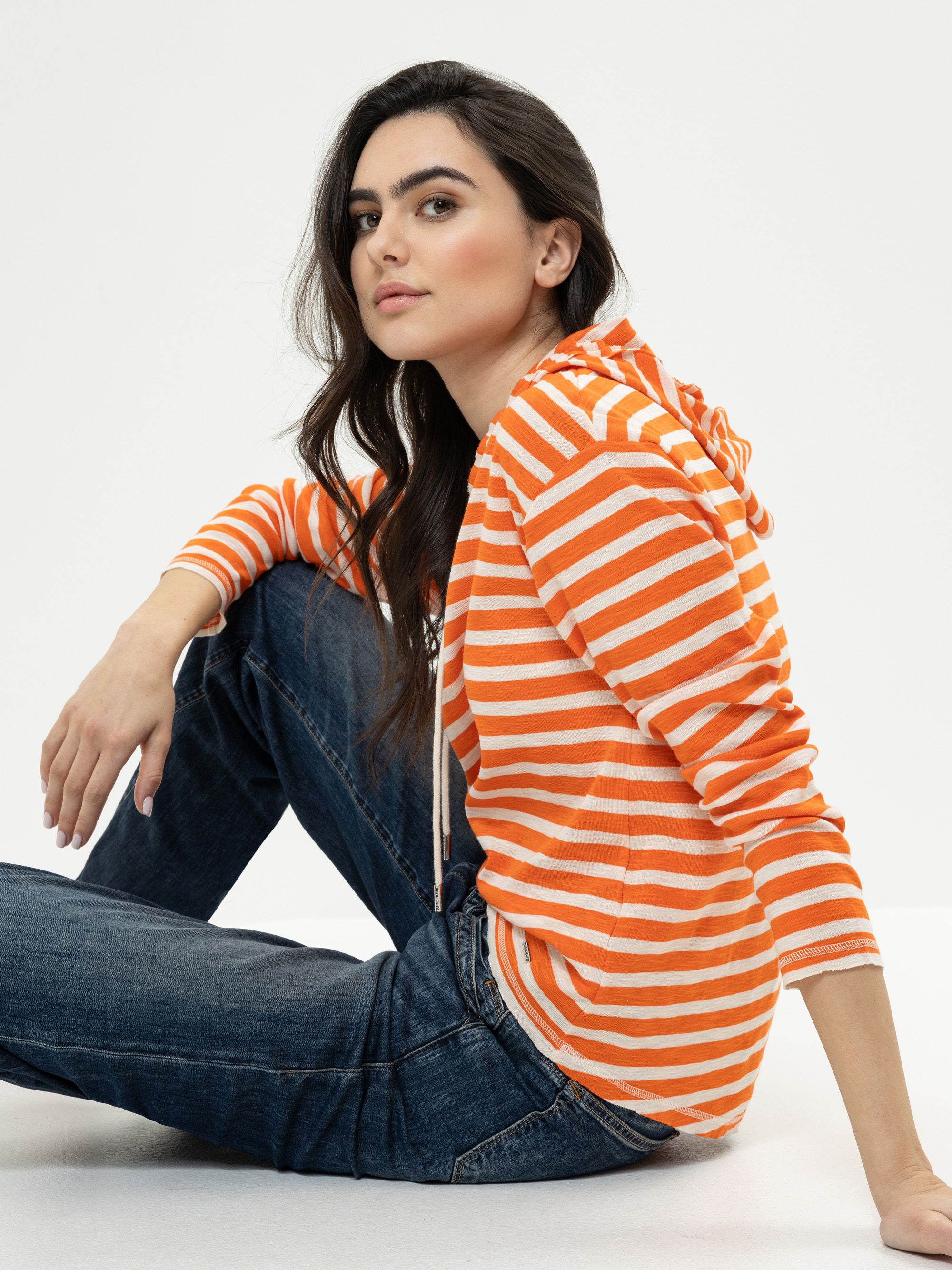 Eine Frau in einem leuchtend orange-weiß gestreiften Kapuzenpulli und blauen Jeans sitzt auf dem Boden und blickt mit neutralem Gesichtsausdruck in die Kamera vor einem schlichten weißen Hintergrund.