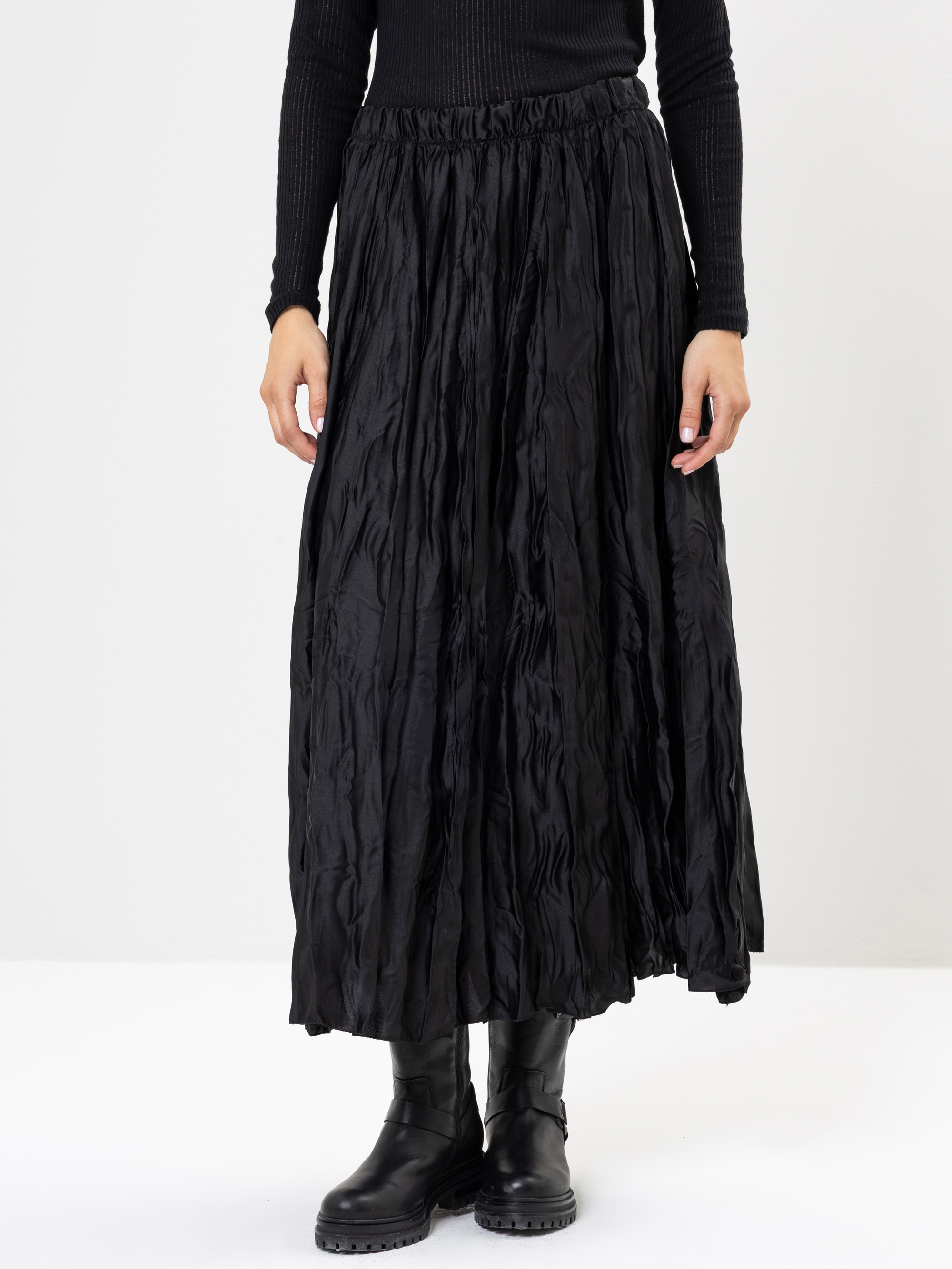 Eine Person, die einen langen schwarzen Faltenrock, ein schwarzes geripptes langärmeliges Oberteil und schwarze Stiefel trägt, steht vor einem schlichten hellen Hintergrund.