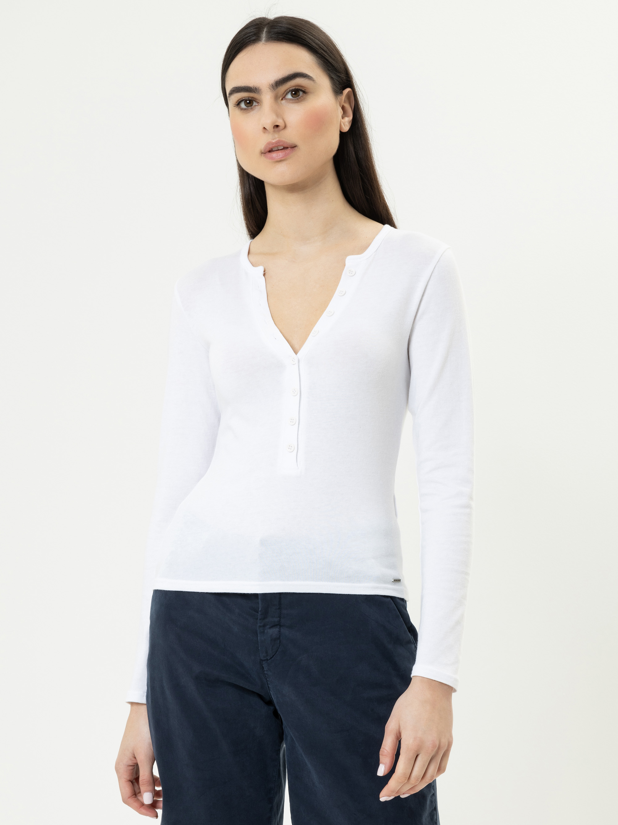 Eine Frau mit glattem dunklem Haar, die ein langärmeliges weißes Hemd und eine dunkle Hose trägt, steht vor einem schlichten hellen Hintergrund und schaut mit neutralem Blick in die Kamera.