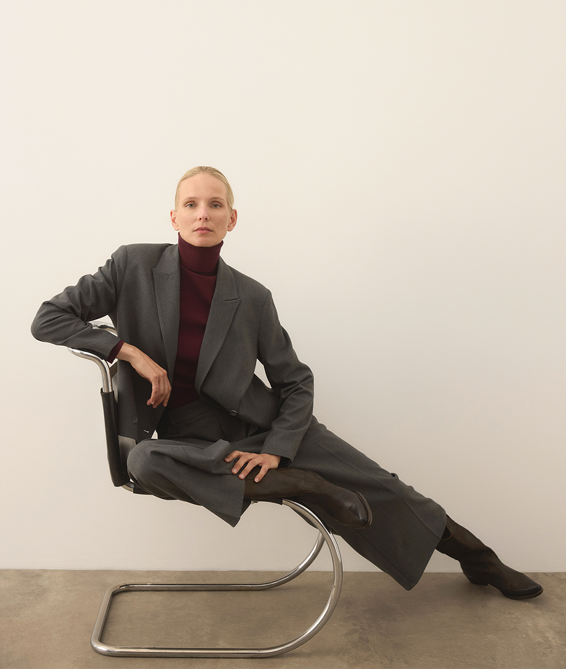 Eine Person mit kurzen blonden Haaren im grauen Anzug und burgunderrotem Rollkragenpullover sitzt selbstbewusst auf einem modernen Metallstuhl, ein Bein ausgestreckt und die Arme entspannt, vor einer schlichten, grauweißen Wand und einem Betonboden.