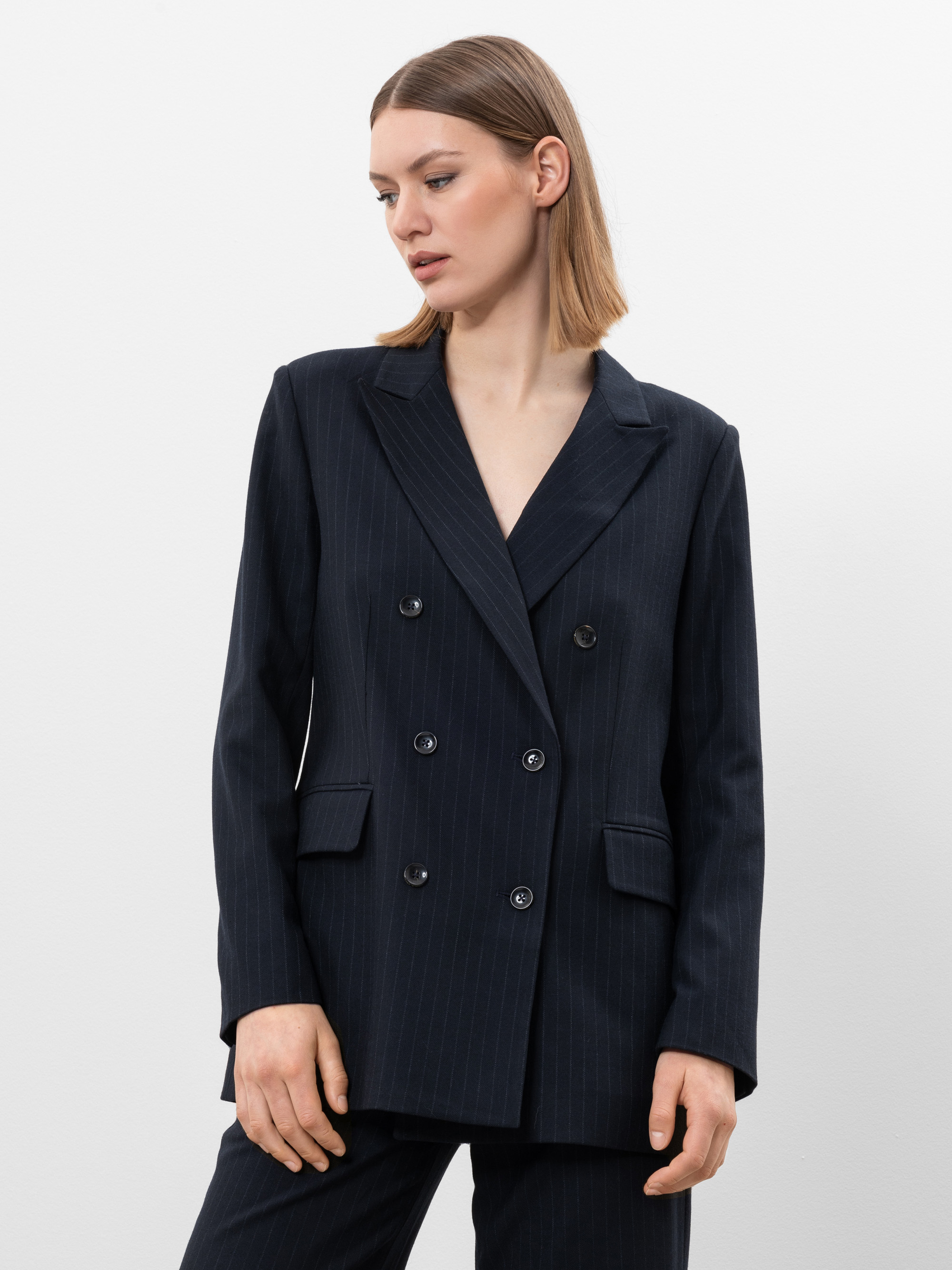 Eine Frau mit glattem, schulterlangem hellbraunem Haar trägt einen dunklen zweireihigen Blazer mit Nadelstreifen und eine dazu passende Hose. Sie steht vor einem schlichten weißen Hintergrund und blickt leicht nach links.