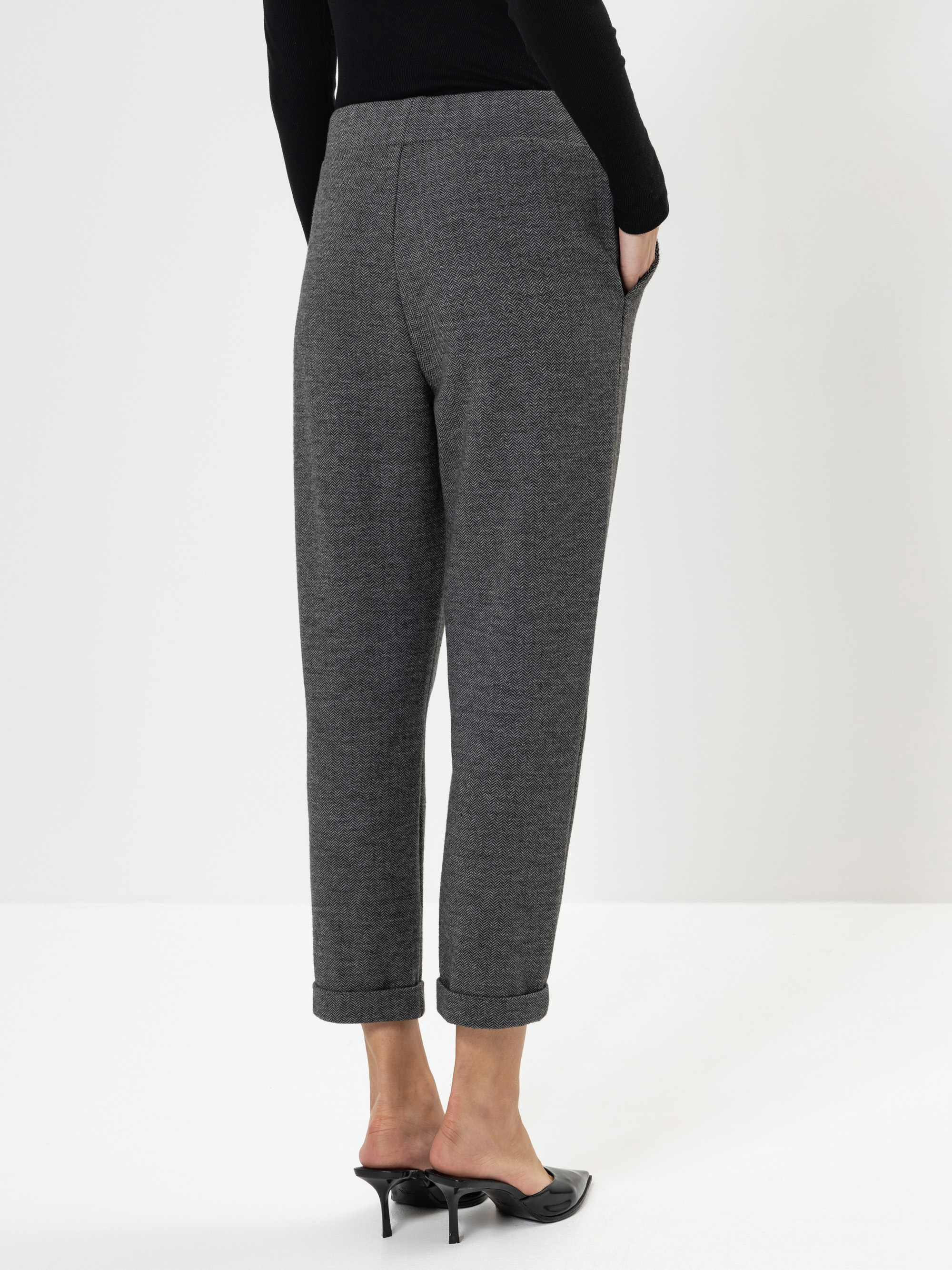 Eine Person, die eine graue, mit Manschetten versehene Hose und schwarze Pantoletten mit Absatz trägt, steht mit den Händen in den Taschen, von der Kamera abgewandt, vor einem schlichten weißen Hintergrund.