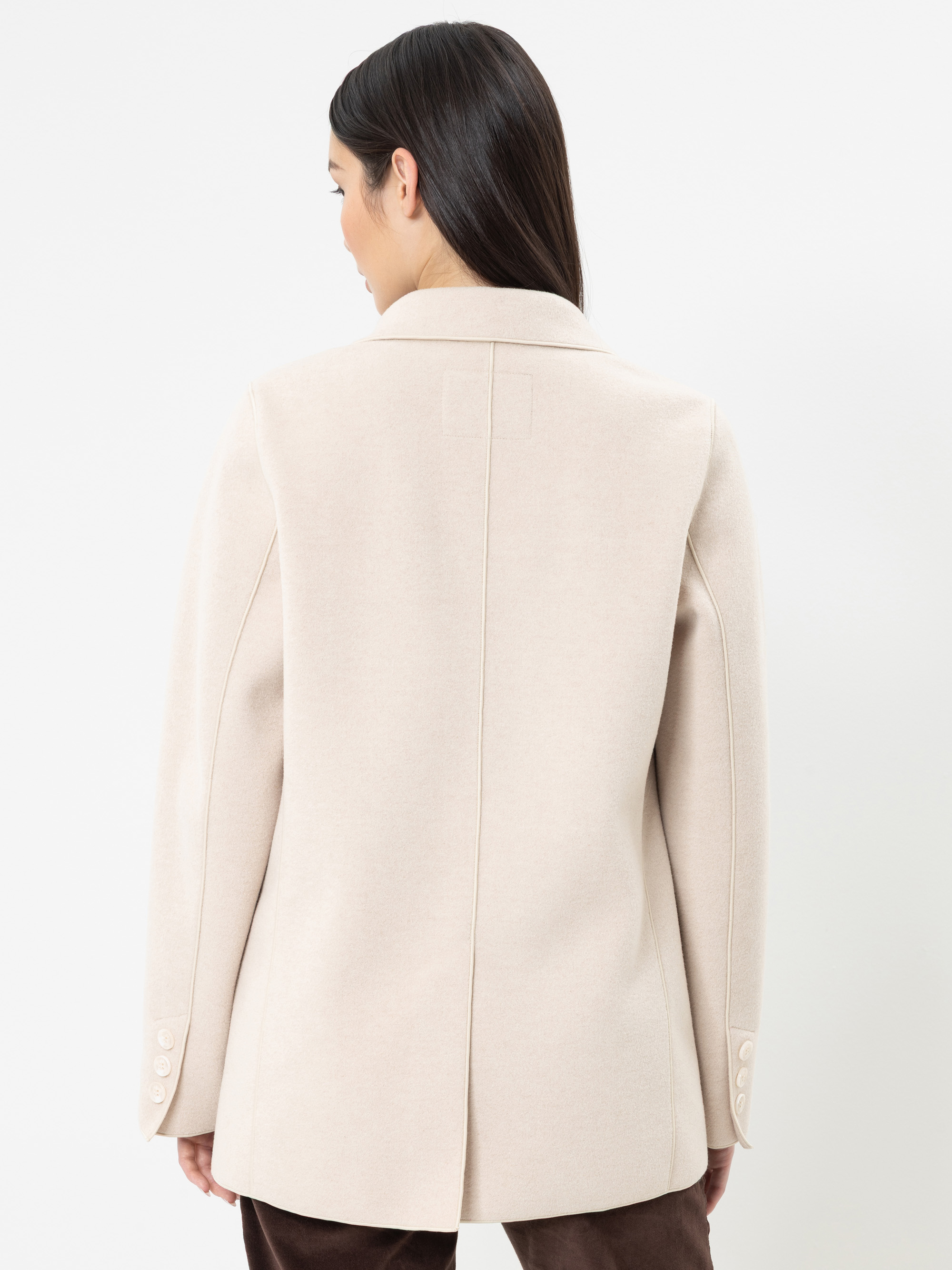 Eine Frau mit langen dunklen Haaren, die einen hellbeigen Blazer und eine braune Hose trägt, ist von hinten vor einem schlichten weißen Hintergrund zu sehen.