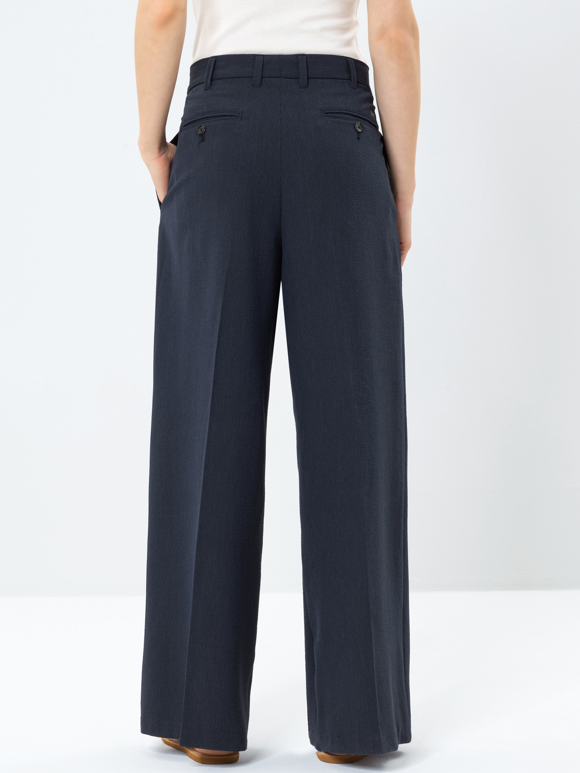 Eine Person, die eine marineblaue Hose mit hoher Taille und weitem Bein trägt, ist von hinten zu sehen, wobei sie die Hände in den Taschen hat. Die Hose hat geknöpfte Gesäßtaschen und wird mit einem weißen Oberteil kombiniert, das in die Hose gestec