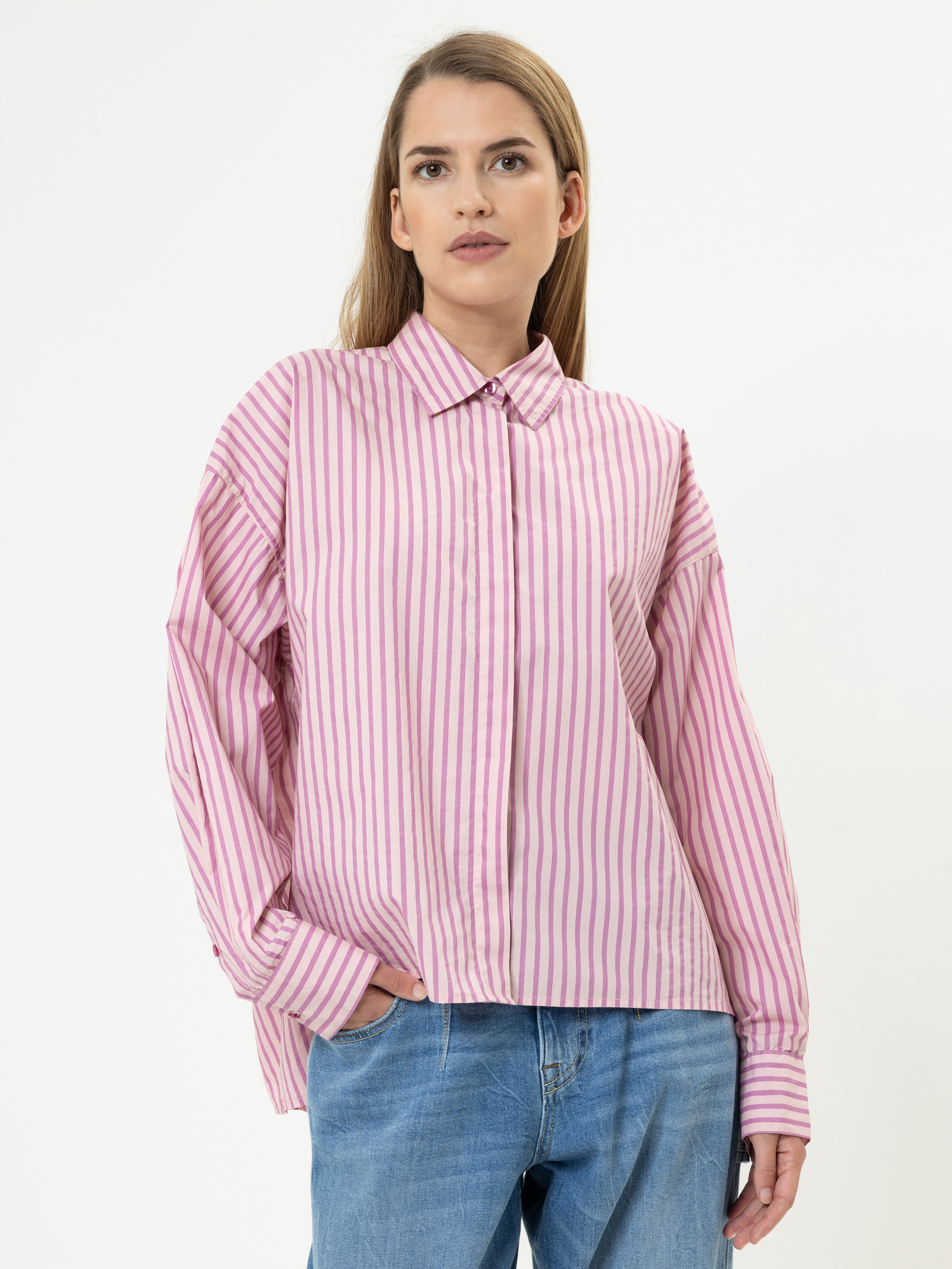 Eine Frau mit glattem, hellbraunem Haar trägt ein locker sitzendes, langärmeliges, rosa-weiß gestreiftes Hemd und blaue Jeans. Sie steht vor einem schlichten weißen Hintergrund und hat eine Hand in der Tasche.