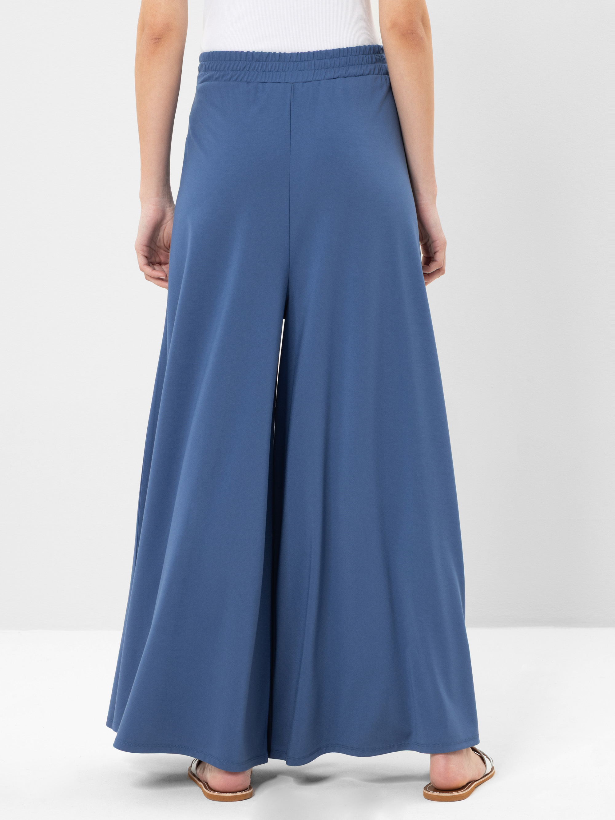 Gezeigt wird eine Person von hinten, die eine weite, blaue Hose und flache Sandalen trägt. Die Hose hat einen elastischen Bund und ist mit einem weißen Oberteil kombiniert. Der Hintergrund ist einfarbig und hell.