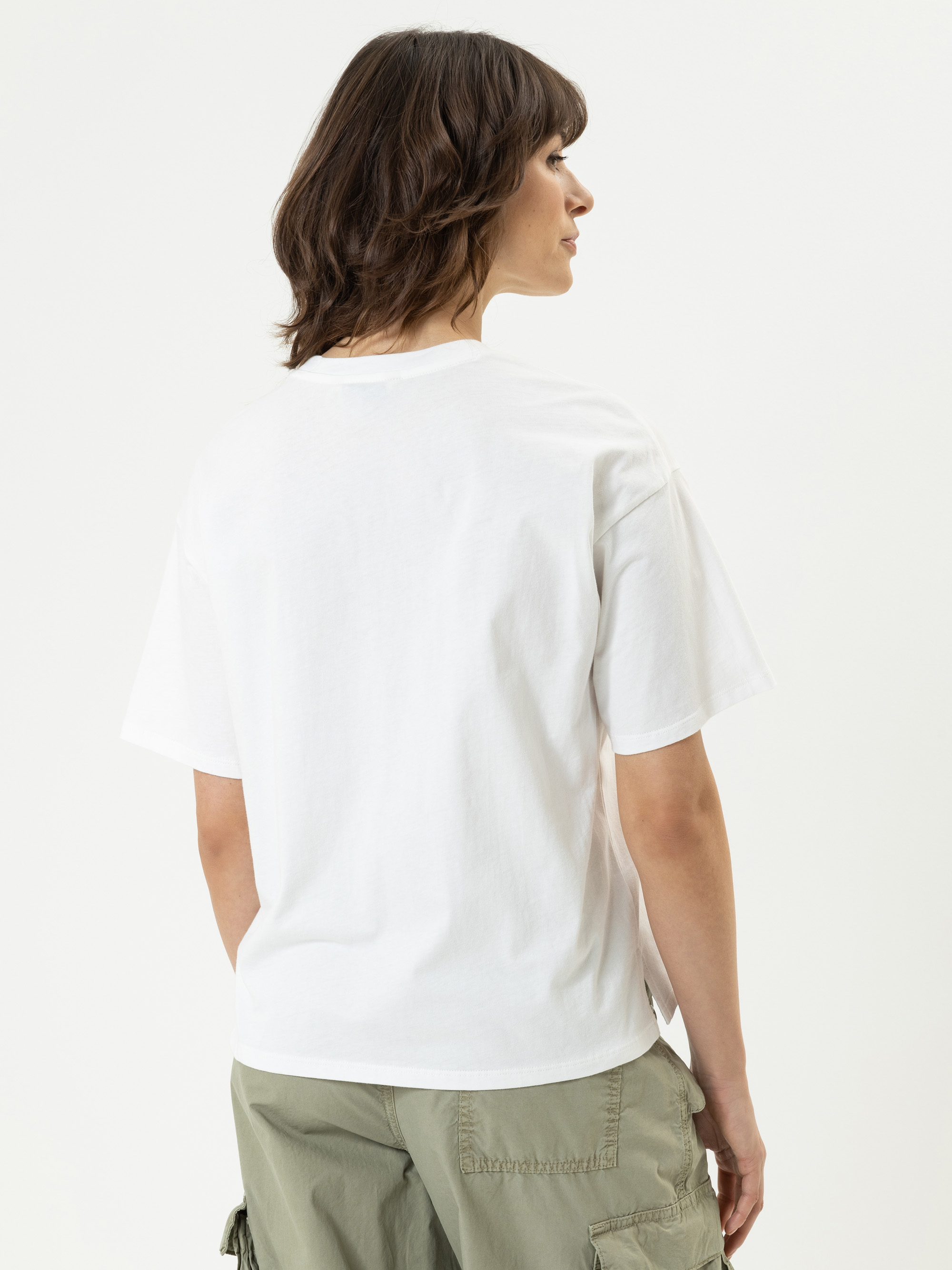 Eine Person mit schulterlangen braunen Haaren, die ein schlichtes weißes T-Shirt und eine hellgrüne Cargohose trägt, steht mit dem Gesicht zur Kamera vor einem weißen Hintergrund.