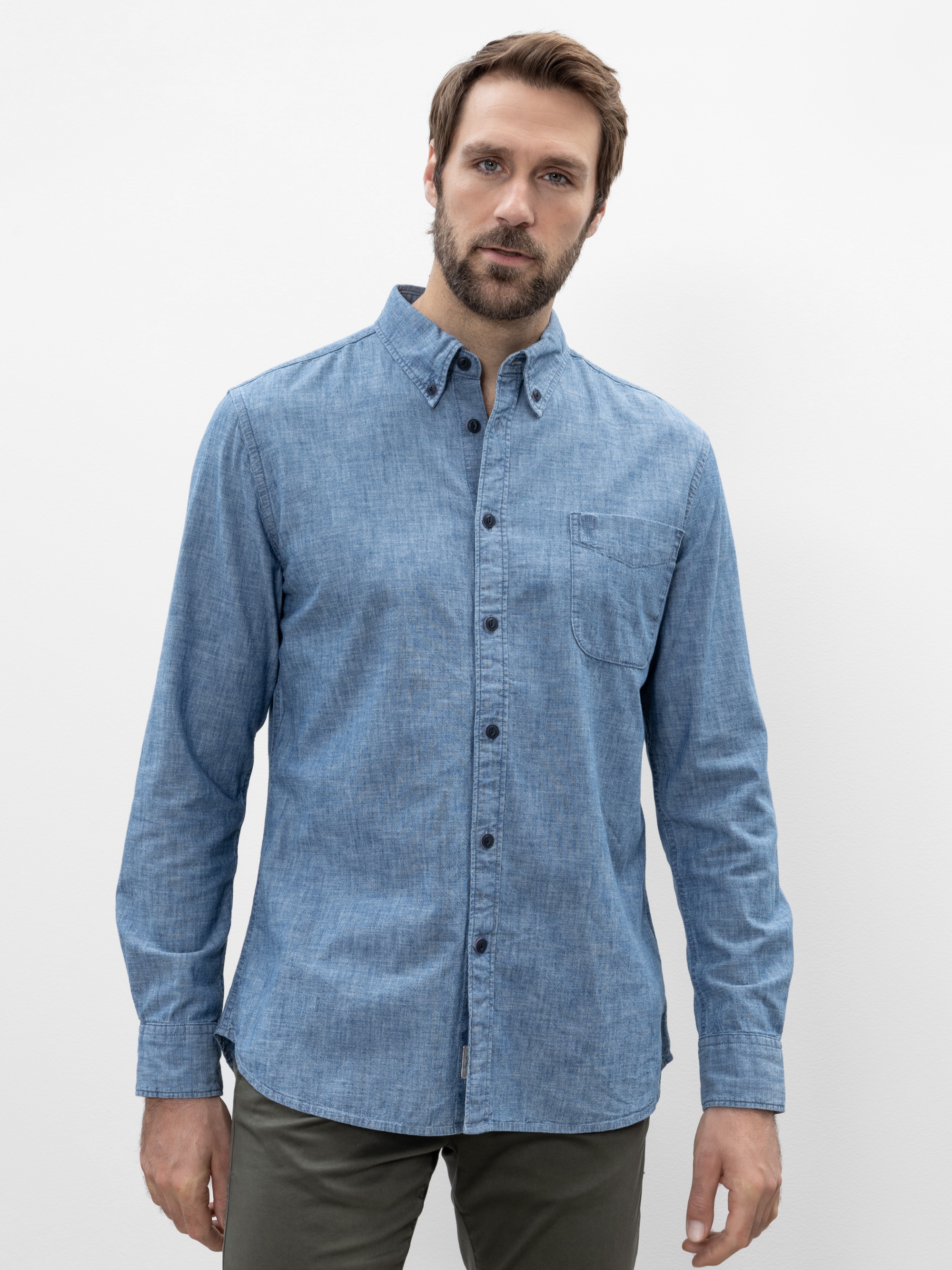 Ein Mann mit kurzen braunen Haaren und Bart trägt ein hellblaues Button-up-Hemd mit Brusttasche und eine dunkle Hose und steht vor einem schlichten weißen Hintergrund.