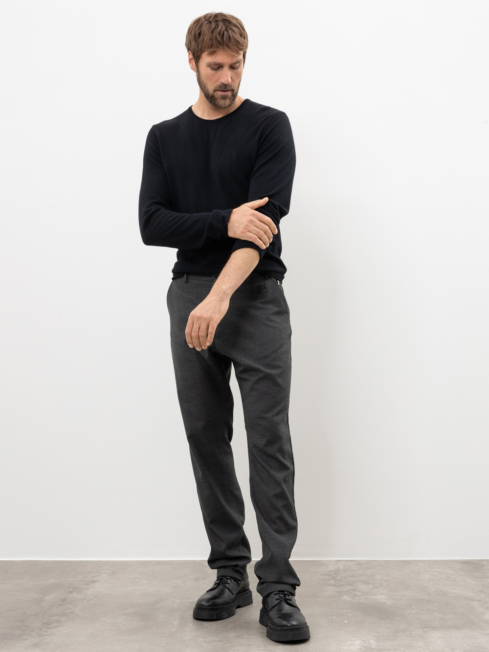 Ein Mann mit kurzen braunen Haaren, der ein schwarzes langärmeliges Hemd, eine dunkelgraue Hose und schwarze Schuhe trägt, steht in einem minimalistischen Raum mit weißer Wand und Betonboden, rückt seinen Ärmel zurecht und schaut nach unten.