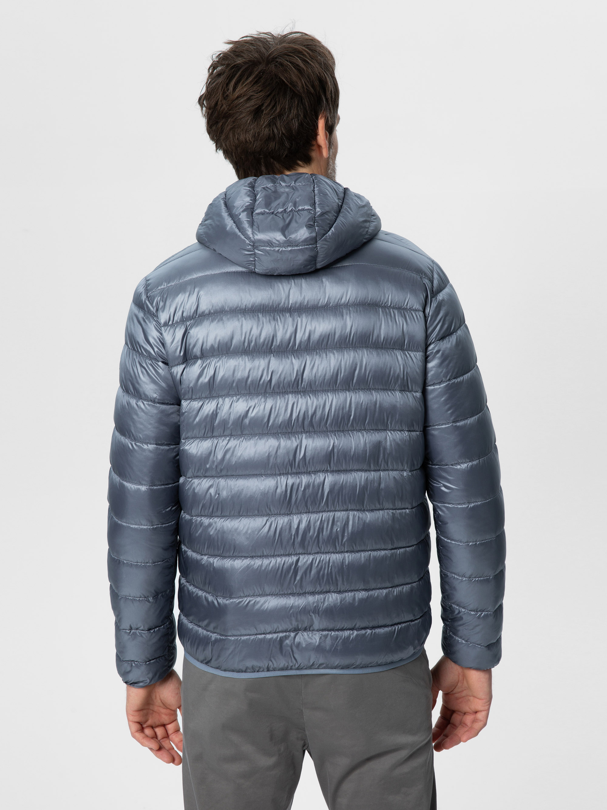 Eine Person mit kurzen braunen Haaren, von hinten gesehen, trägt eine glänzende graue Pufferjacke mit Kapuze und eine graue Hose und steht vor einem schlichten hellen Hintergrund.