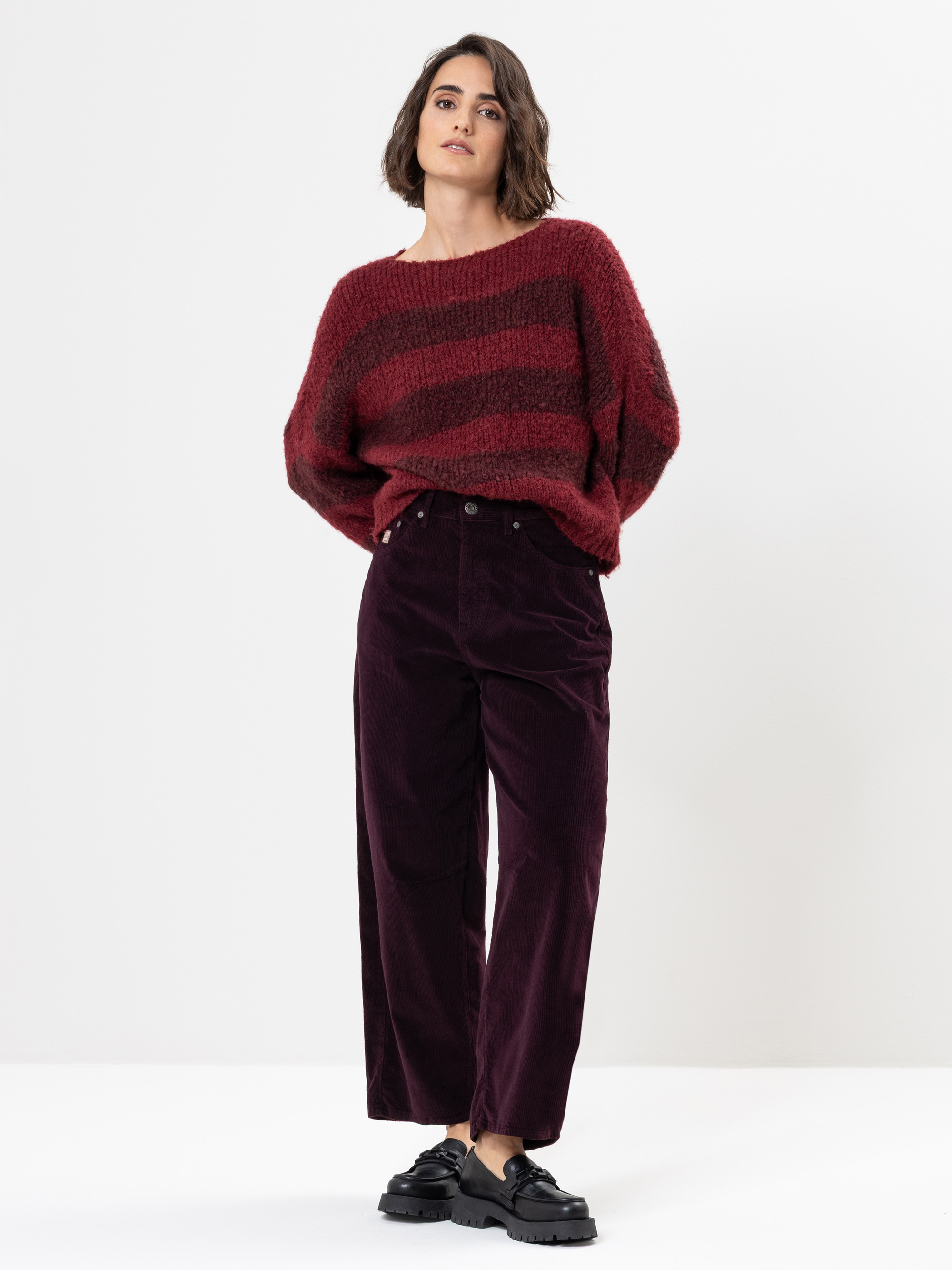 Eine Frau steht vor einem weißen Hintergrund und trägt einen lockeren, rot und burgunderrot gestreiften Pullover, eine dunkle Hose mit weitem Bein und schwarze, klobige Schuhe. Sie hat die Hände hinter dem Rücken verschränkt und blickt leicht nach ob