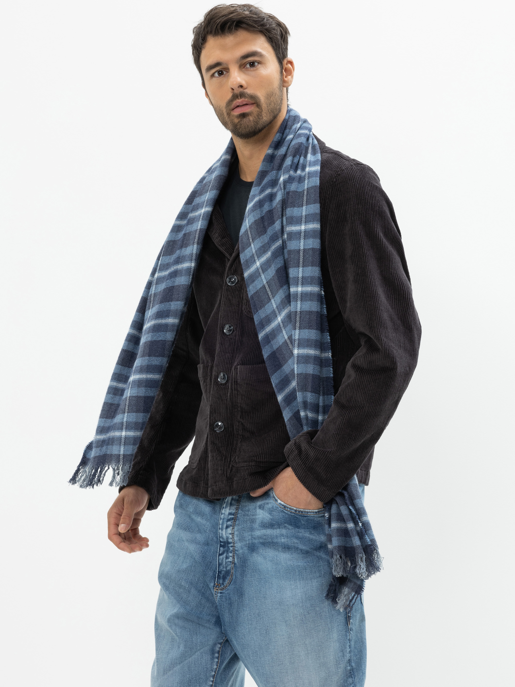 Ein Mann in schwarzer Jacke, blauer Jeans und blau-kariertem Schal steht vor einem schlichten weißen Hintergrund und blickt selbstbewusst in die Kamera, eine Hand in der Tasche.