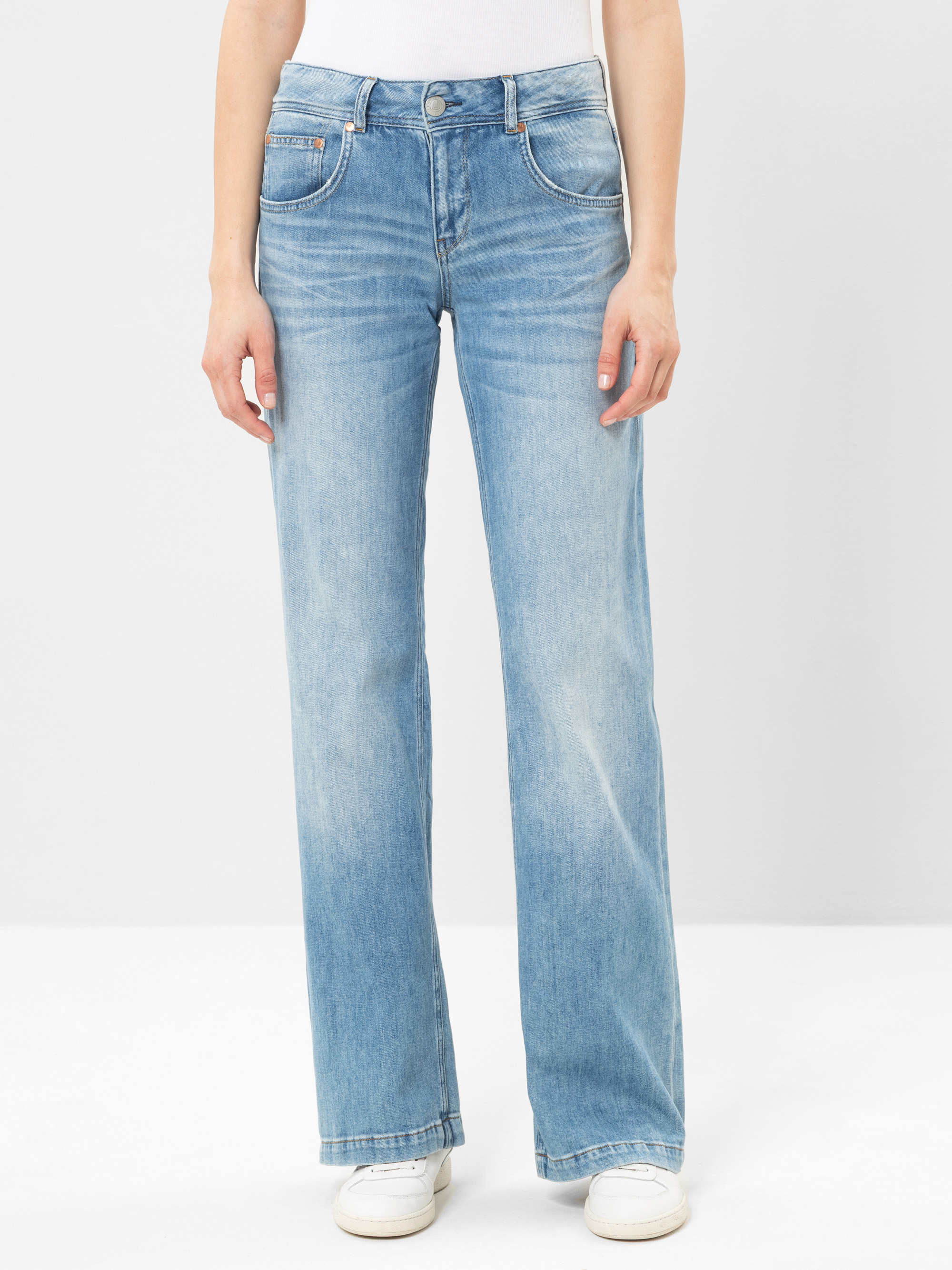 Eine Person, die hellblaue Jeans mit hoher Taille und geradem Bein mit leichten Ausbleichungen, ein weißes Oberteil und weiße Turnschuhe trägt, steht vor einem schlichten hellen Hintergrund.
