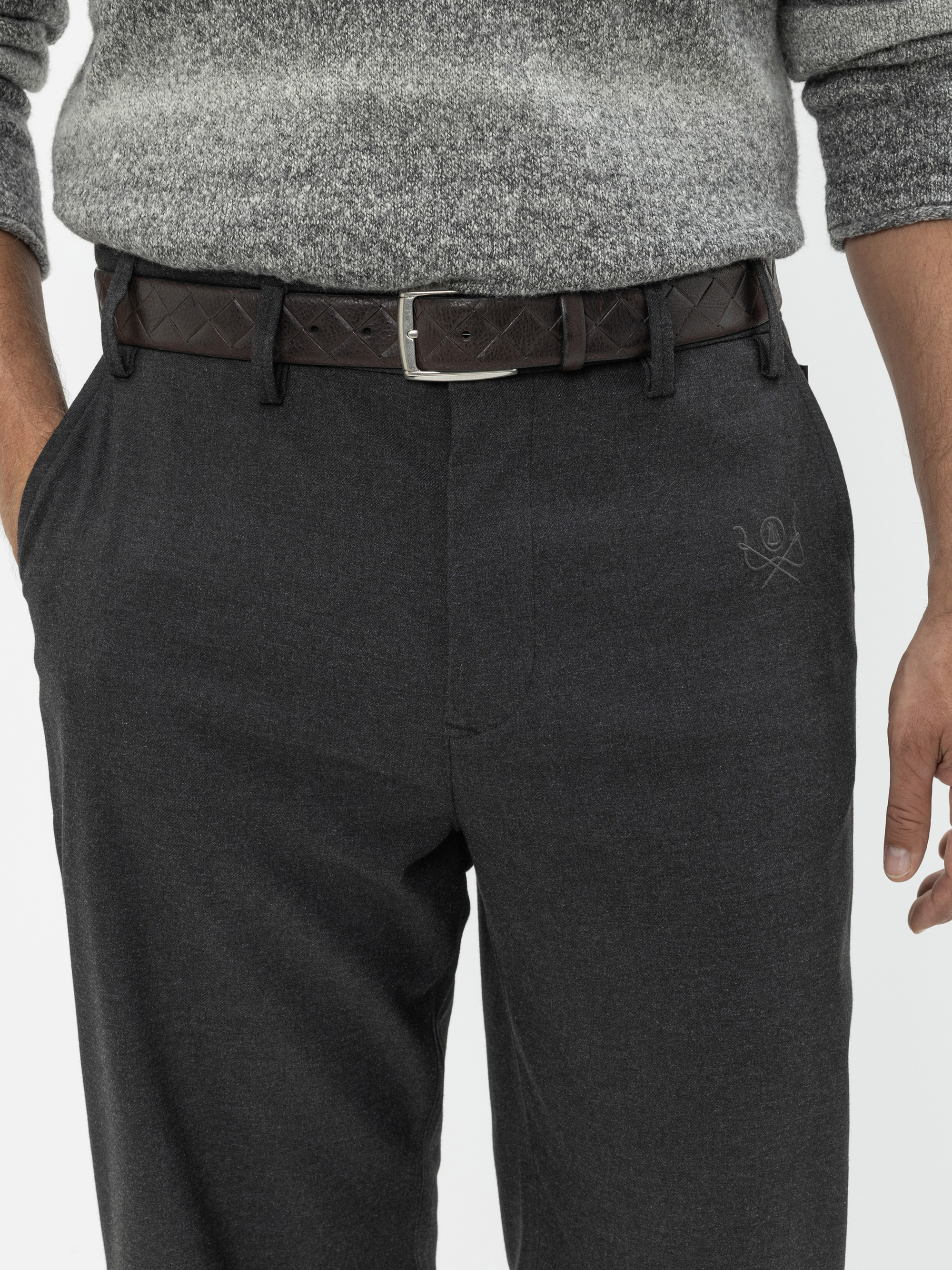 Eine Person trägt eine dunkelgraue Anzughose, einen braunen Flechtgürtel und einen grauen Strickpullover. Die Hände befinden sich in den Hosentaschen, und nur der Mittelteil ist vor einem schlichten Hintergrund zu sehen.