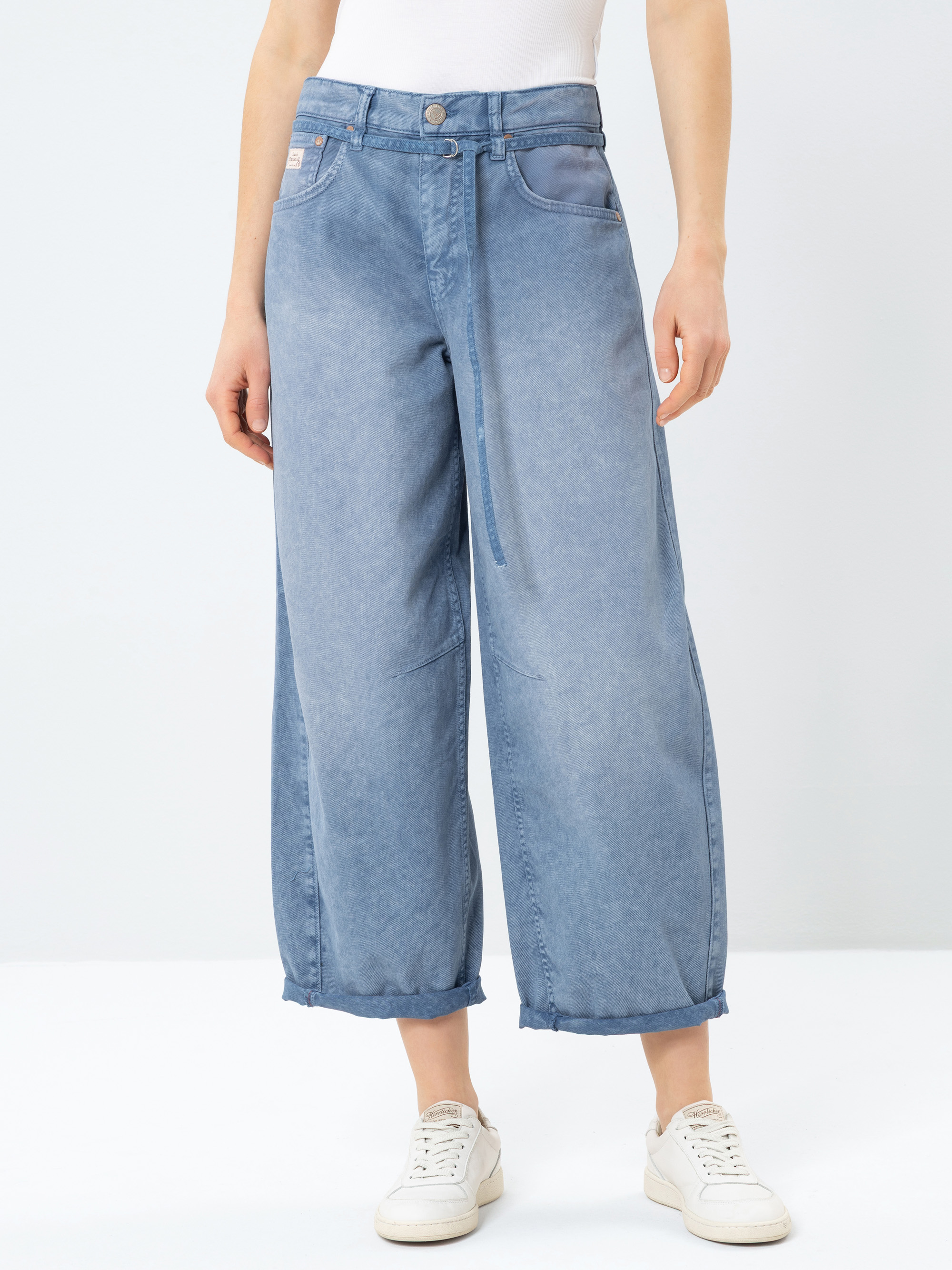 Eine Person, die hellblaue, weit geschnittene Jeans mit gekrempelten Säumen, ein weißes Oberteil und weiße Turnschuhe trägt, steht vor einem schlichten hellen Hintergrund.