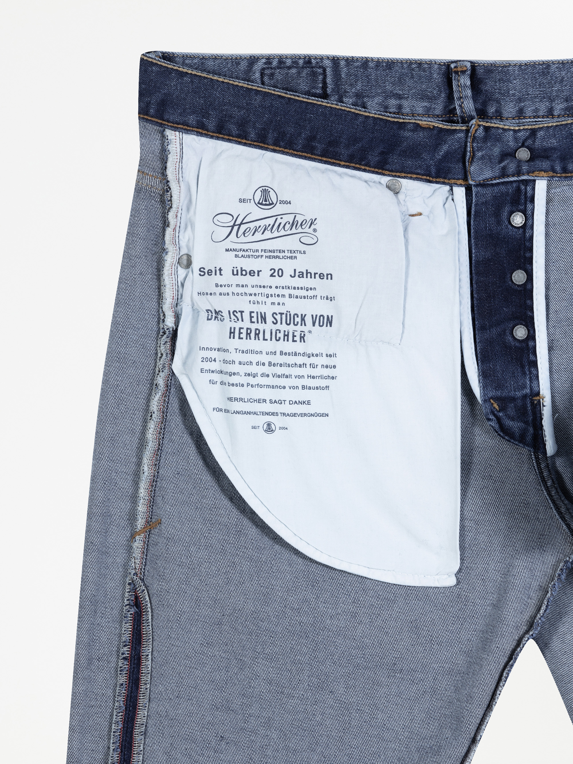 Nahaufnahme der Innenseite einer blauen Jeans, die ein weißes Taschenfutter mit aufgedrucktem Text und Markeninformationen in deutscher Sprache, Metallknöpfe und sichtbare Nähte zeigt.