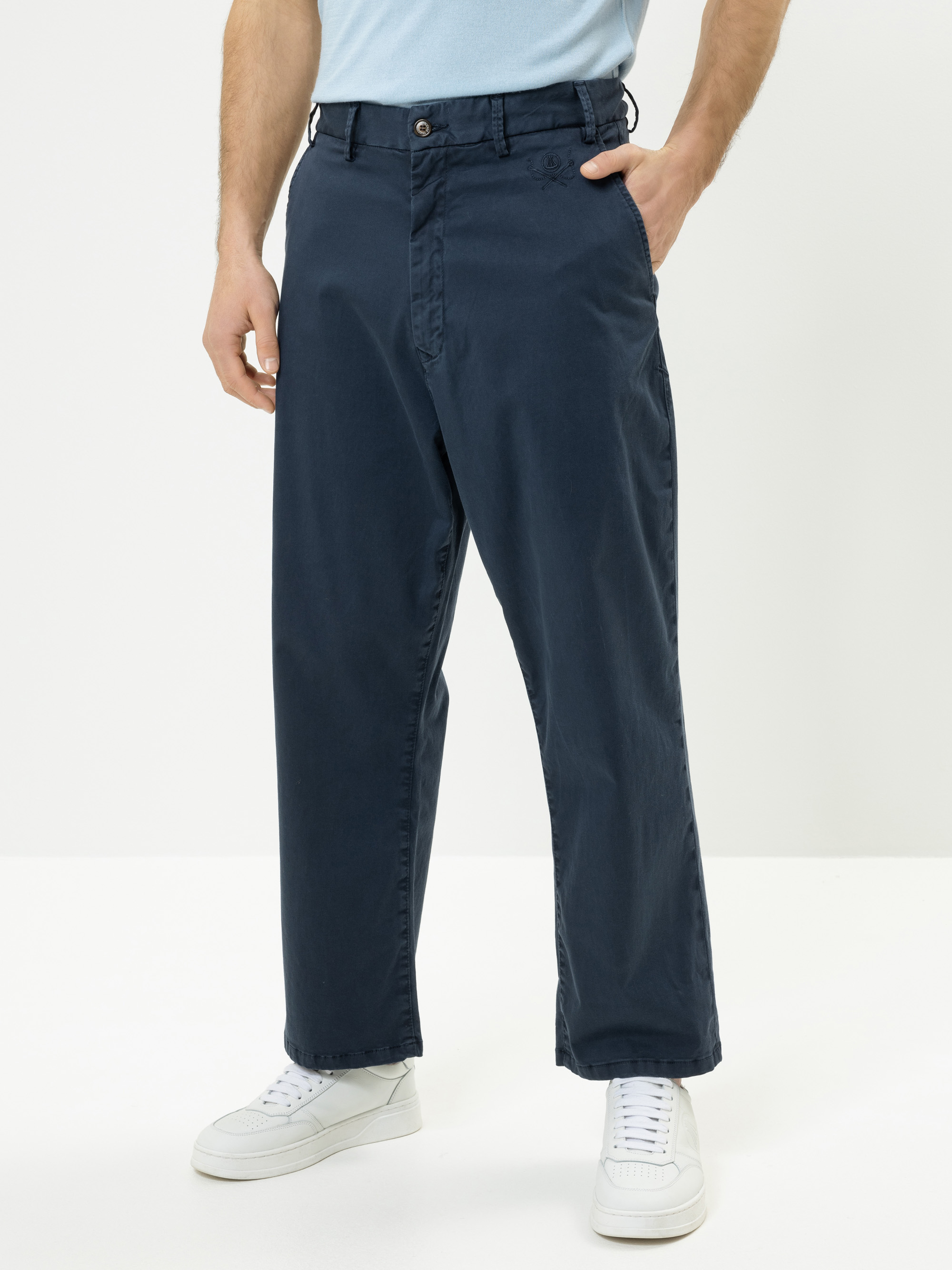 Eine Person, die eine hochgezogene, locker sitzende marineblaue Hose, ein hellblaues Hemd und weiße Turnschuhe trägt, steht mit einer Hand in der Tasche vor einem schlichten weißen Hintergrund.