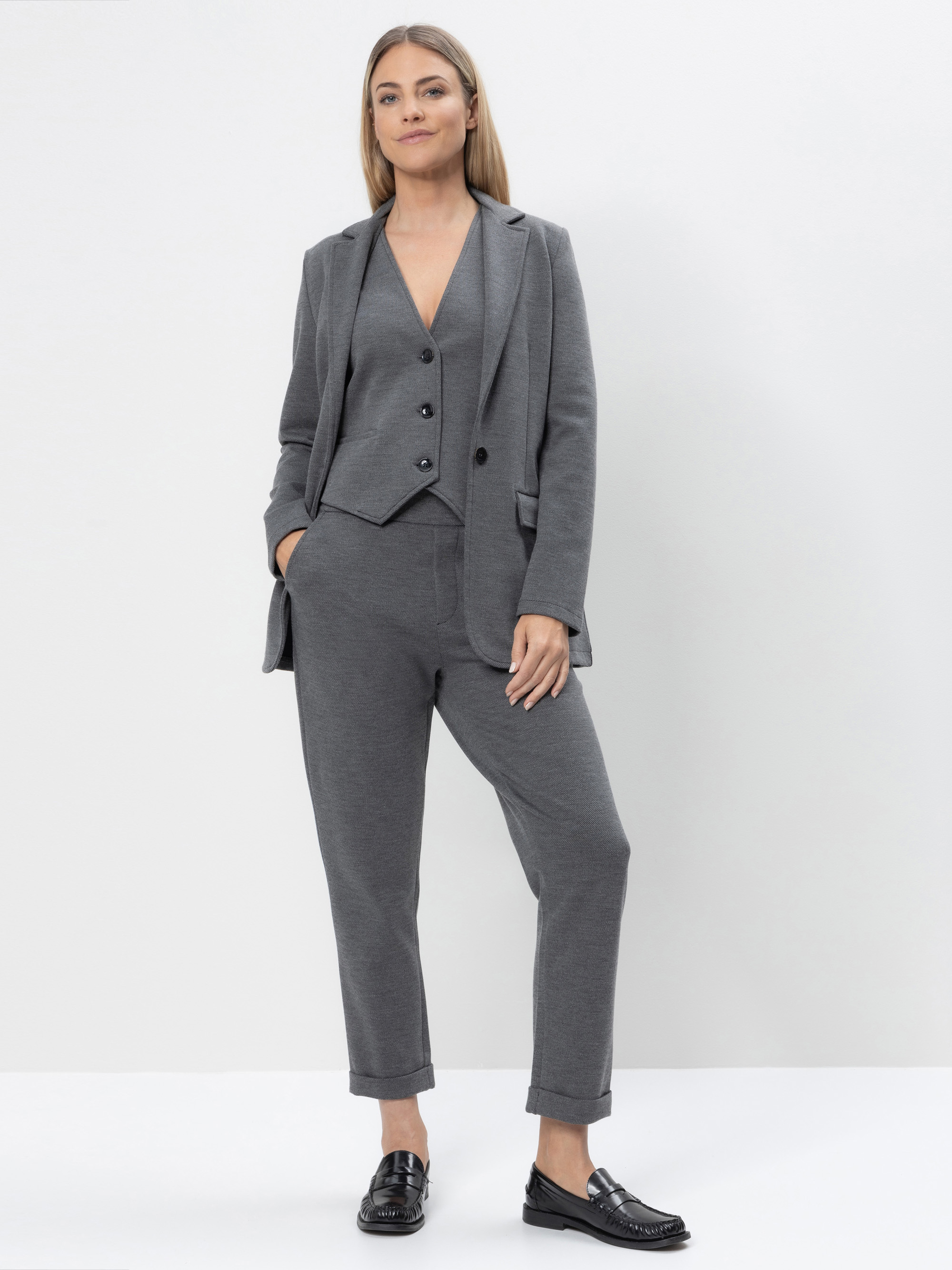 Eine Frau steht vor einem weißen Hintergrund und trägt einen grauen Blazer, eine passende Weste und eine Hose. Sie hat die Hände in den Hosentaschen und trägt schwarze Slipper. Sie wirkt entspannt und selbstbewusst.
