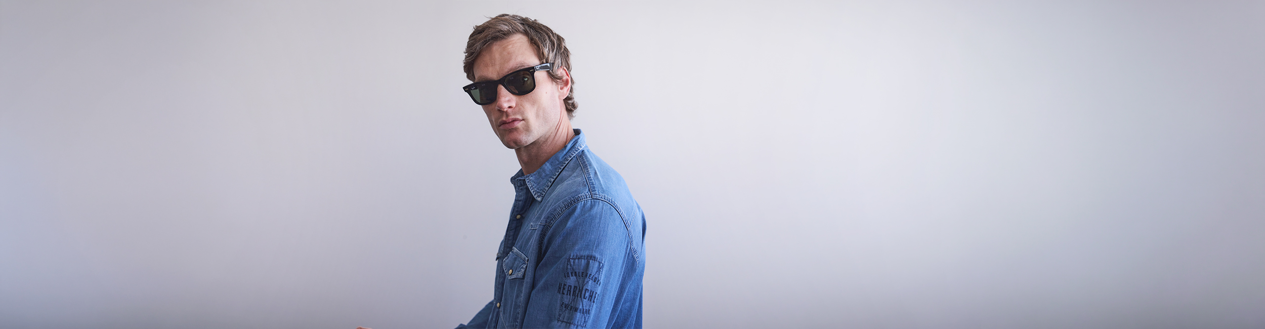 Eine Person mit schwarzer Sonnenbrille und einem blauen Jeanshemd steht vor einem schlichten, hellen Hintergrund und blickt leicht zur Seite.