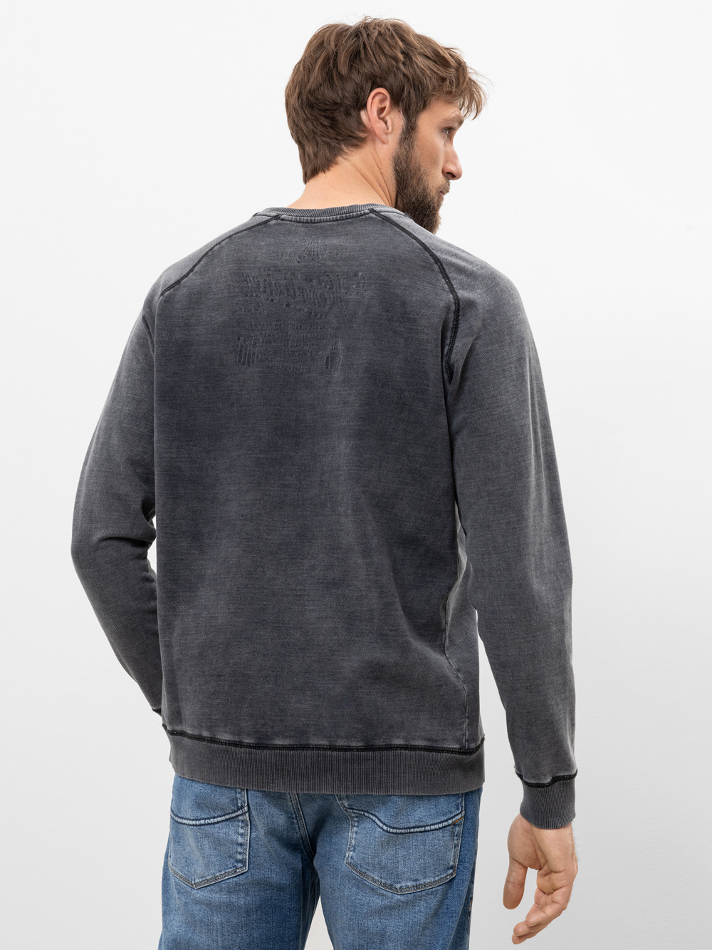 Ein Mann mit braunen Haaren und einem Bart trägt ein verblichenes schwarzes Sweatshirt und blaue Jeans. Er steht mit dem Rücken zur Kamera vor einem schlichten weißen Hintergrund.