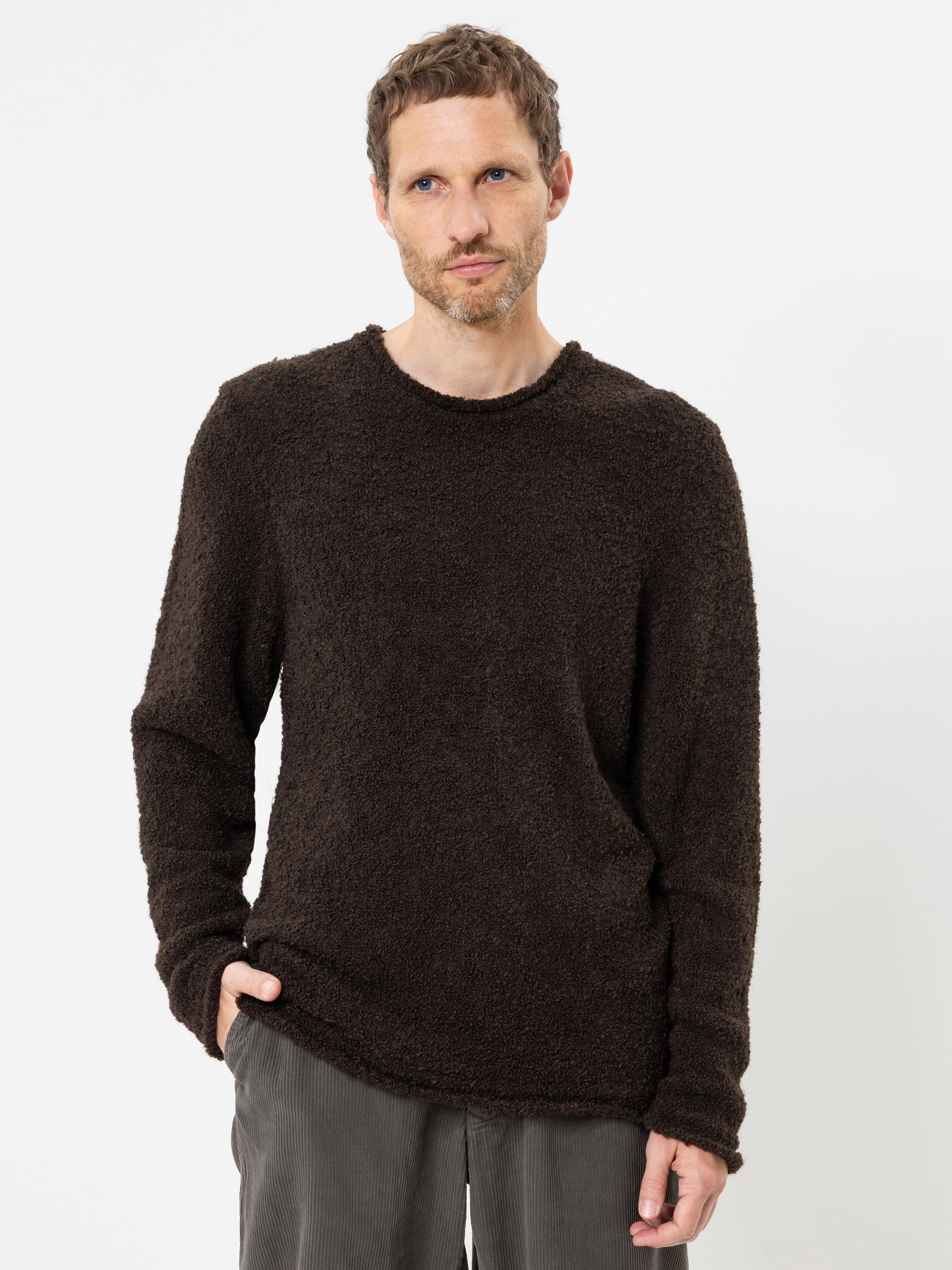 Ein Mann mit kurzen, hellbraunen Haaren und leichtem Bartwuchs trägt einen dunkelbraunen, strukturierten Pullover und eine graue Hose; er steht vor einem schlichten weißen Hintergrund und hat eine Hand in der Hosentasche.