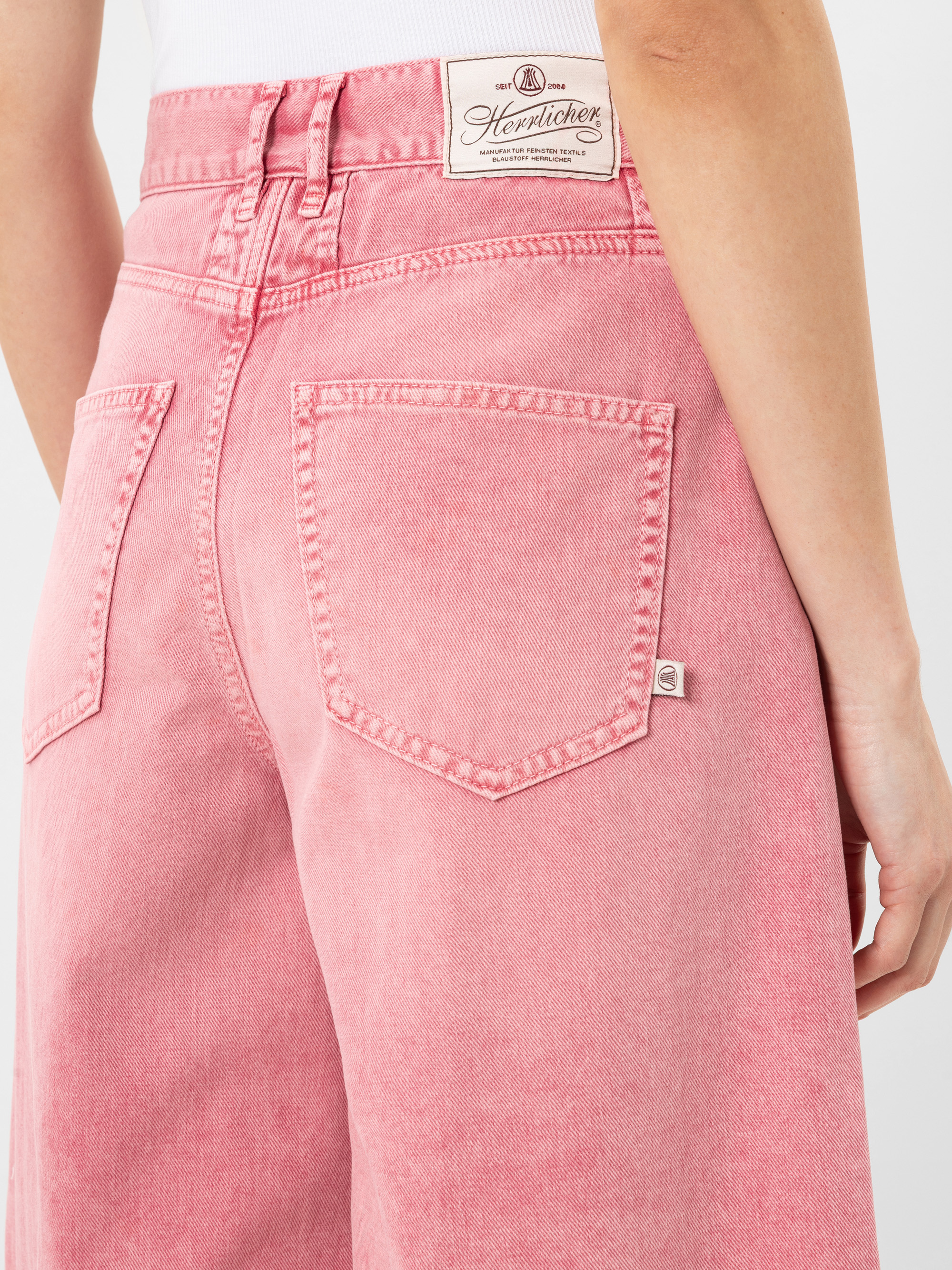 Nahaufnahme einer Person, die eine hochtaillierte, hellrosa Jeans mit großen Gesäßtaschen und einem weißen Markenetikett am Bund trägt. Die Person trägt außerdem ein weißes Oberteil.