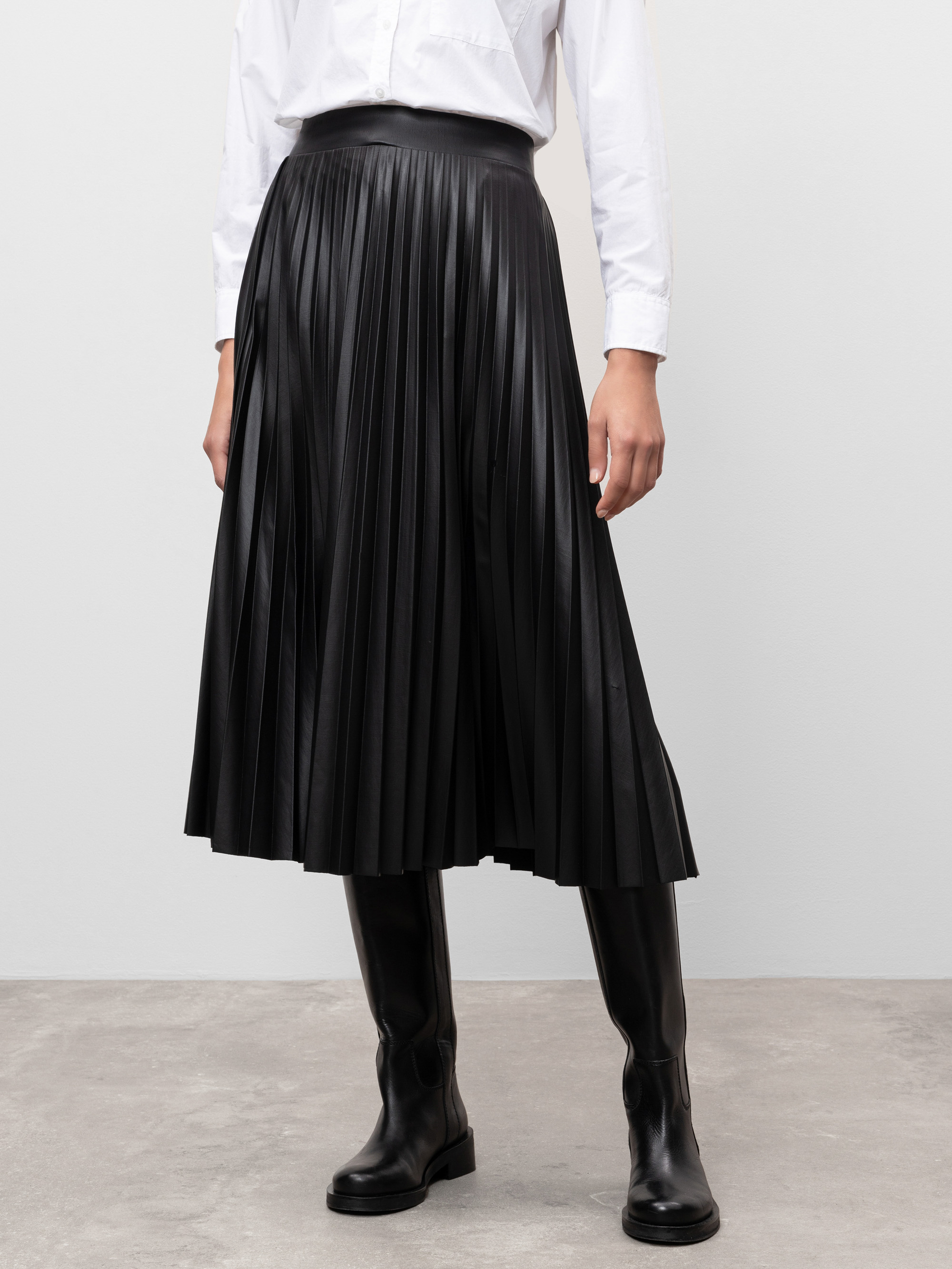 Eine Person, die ein weißes langärmeliges Hemd, einen schwarzen plissierten Midirock und schwarze kniehohe Stiefel trägt, steht auf einem grauen Boden vor einem schlichten hellen Hintergrund. Das Gesicht der Person ist nicht sichtbar.
