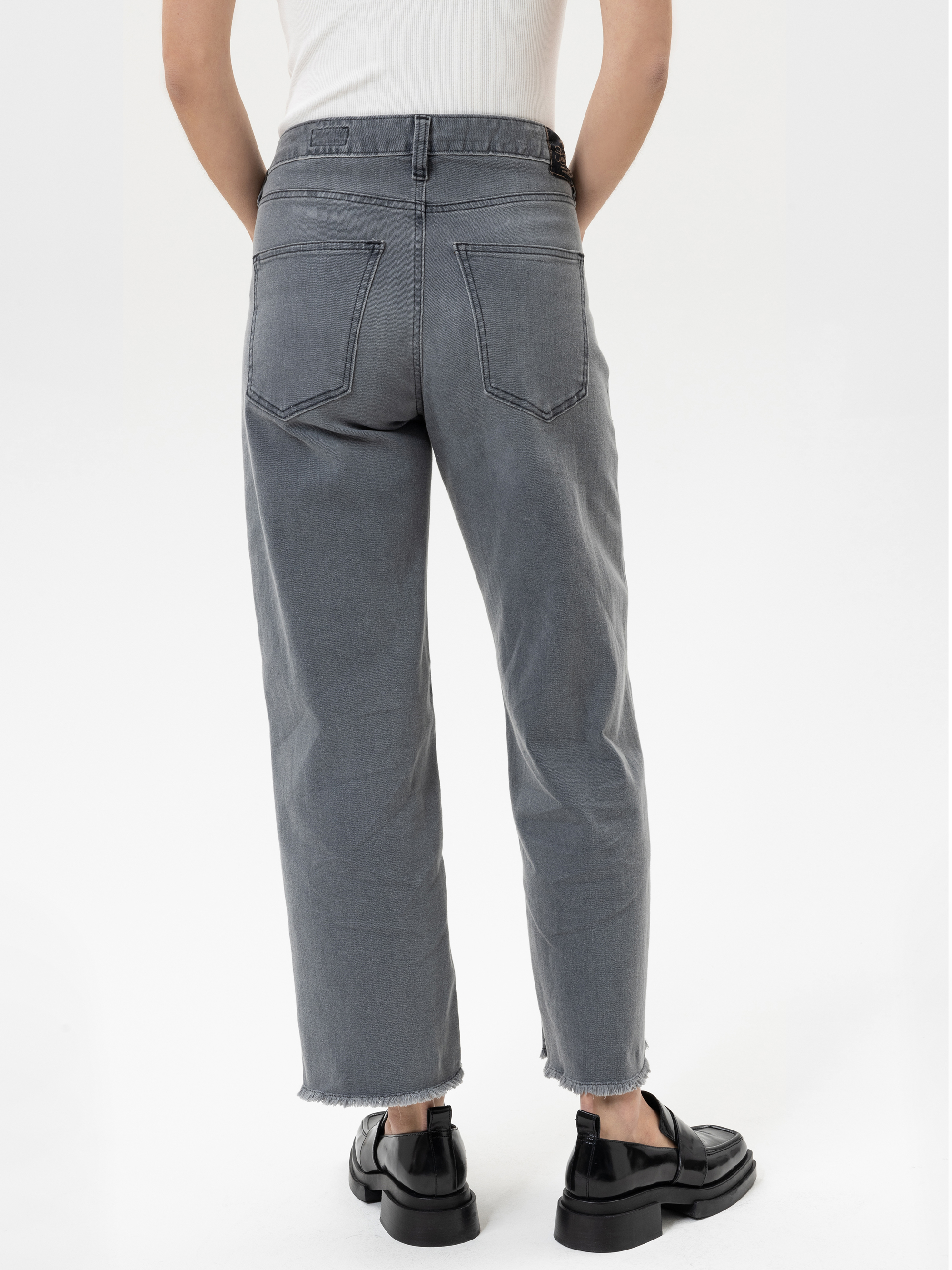 Person, die eine graue Jeans mit hoher Taille und geradem Bein, rohem Saum und schwarzen Schuhen trägt, dargestellt von hinten vor einem einfarbig weißen Hintergrund.