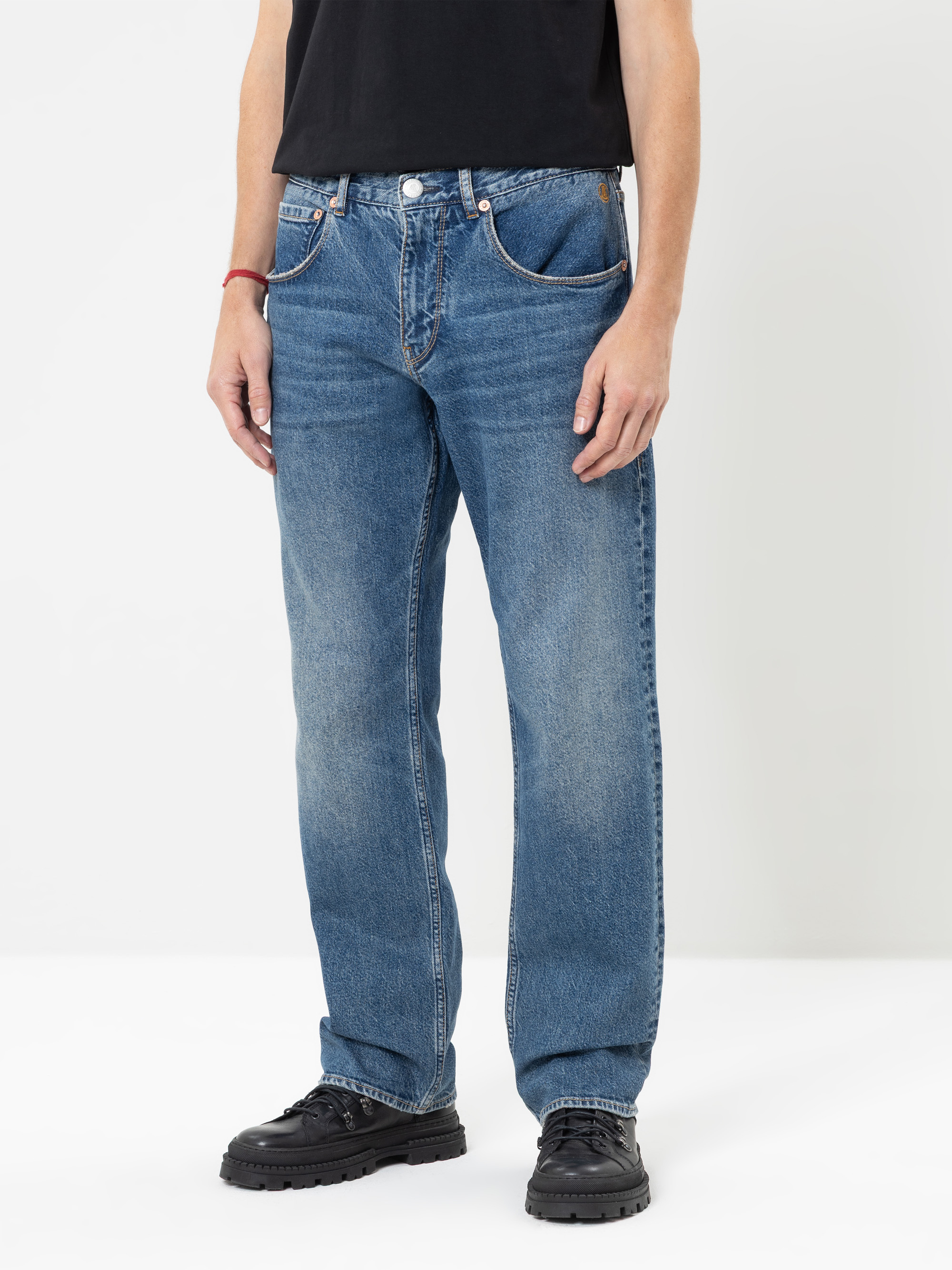 Eine Person in einer locker sitzenden blauen Jeans, einem schwarzen T-Shirt und schwarzen Schuhen steht vor einem schlichten weißen Hintergrund. Zu sehen ist nur die untere Körperhälfte. Die Jeans hat ein klassisches Fünf-Taschen-Design und einen verb