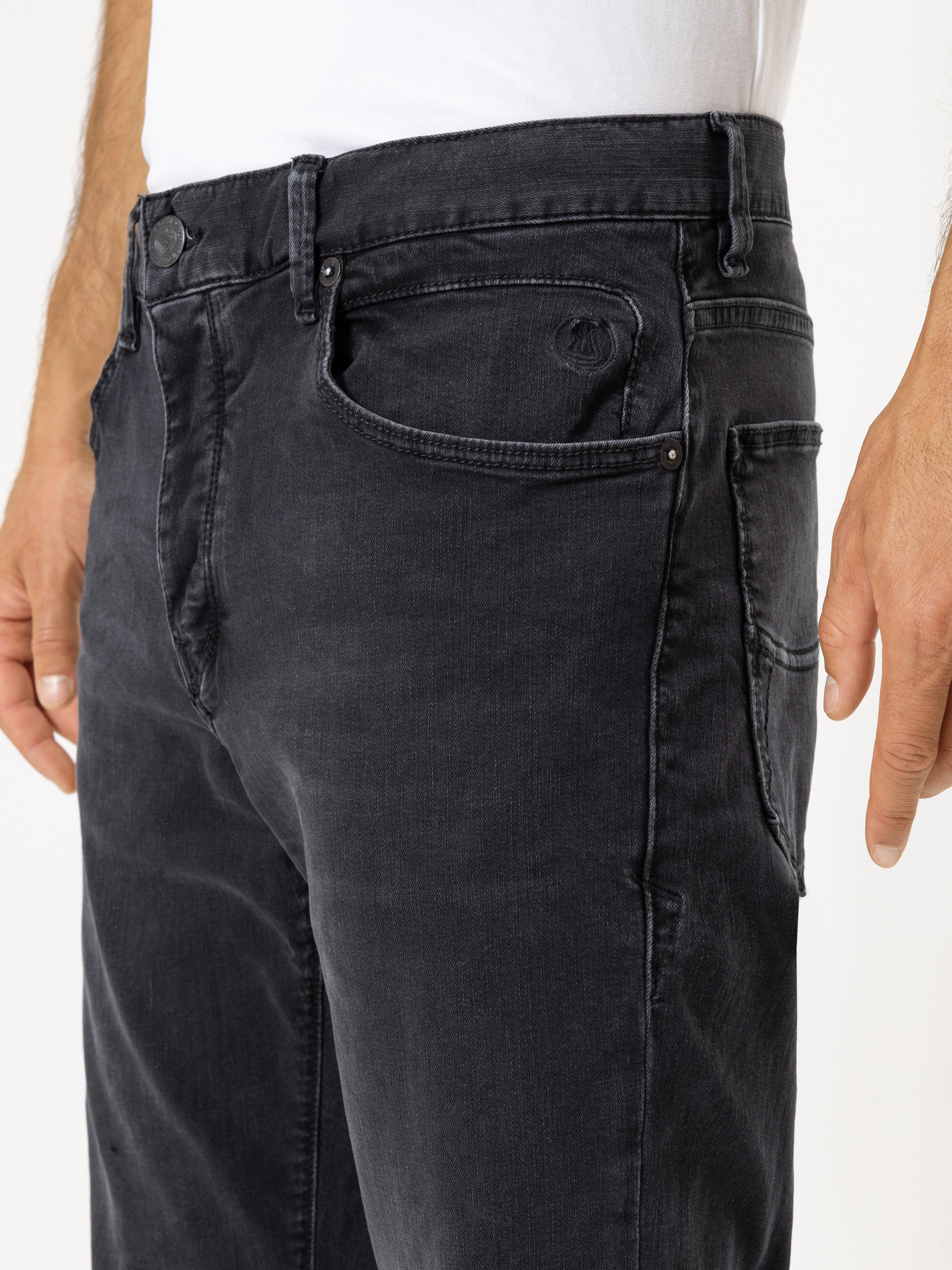 Nahaufnahme einer Person, die eine schwarze Jeans und ein weißes Hemd trägt. Der Fokus liegt auf der Vorderseite und der Seite der Jeans, wobei die Taschen und die Stoffstruktur hervorgehoben werden. Die Hände der Person liegen entspannt an ihren Seite