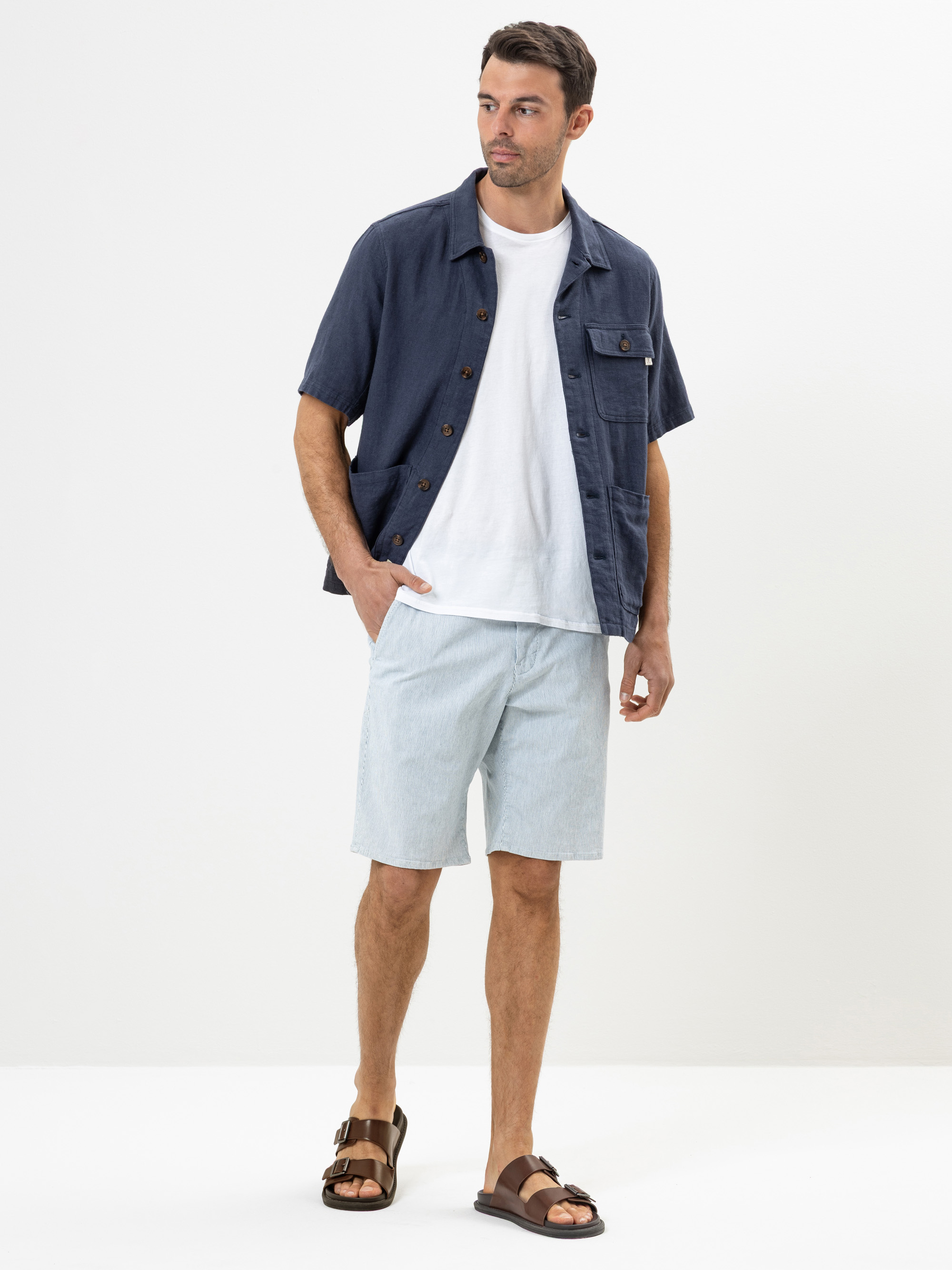 Ein Mann steht vor einem weißen Hintergrund und trägt ein marineblaues kurzärmeliges Hemd über einem weißen T-Shirt, hellblaue Shorts und braune Sandalen. Er blickt leicht zur Seite und hat eine Hand in der Tasche.