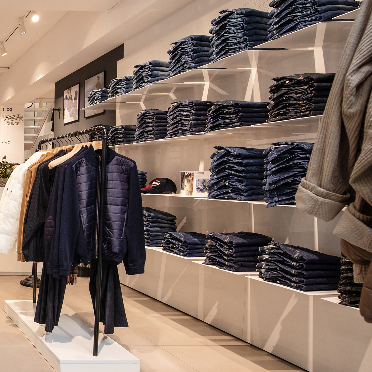 In einem Bekleidungsgeschäft stapeln sich sauber gefaltete Bluejeans in weißen Regalen. Jacken und Hemden hängen in einem Regal im Vordergrund. Der Laden ist modern und übersichtlich gestaltet, Wände und Boden sind in neutralen Tönen gehalten.
