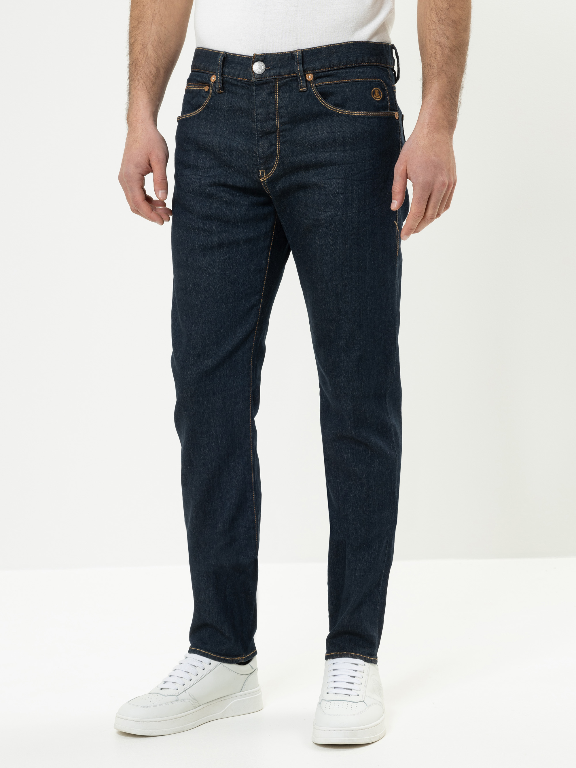 Eine Person steht in dunkelblauen Jeans mit orangefarbenen Nähten, einem weißen T-Shirt und weißen Turnschuhen vor einem schlichten weißen Hintergrund. Das Bild ist so beschnitten, dass nur die untere Hälfte des Körpers zu sehen ist.