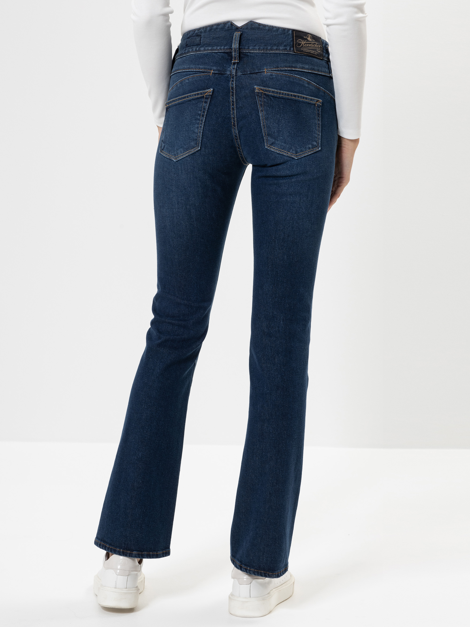 Eine Person, die dunkelblaue Bootcut-Jeans und weiße Turnschuhe trägt, steht von der Kamera weg. Die Jeans hat Gesäßtaschen und ist mit einem weißen langärmeligen Oberteil kombiniert. Der Hintergrund ist einfarbig weiß.