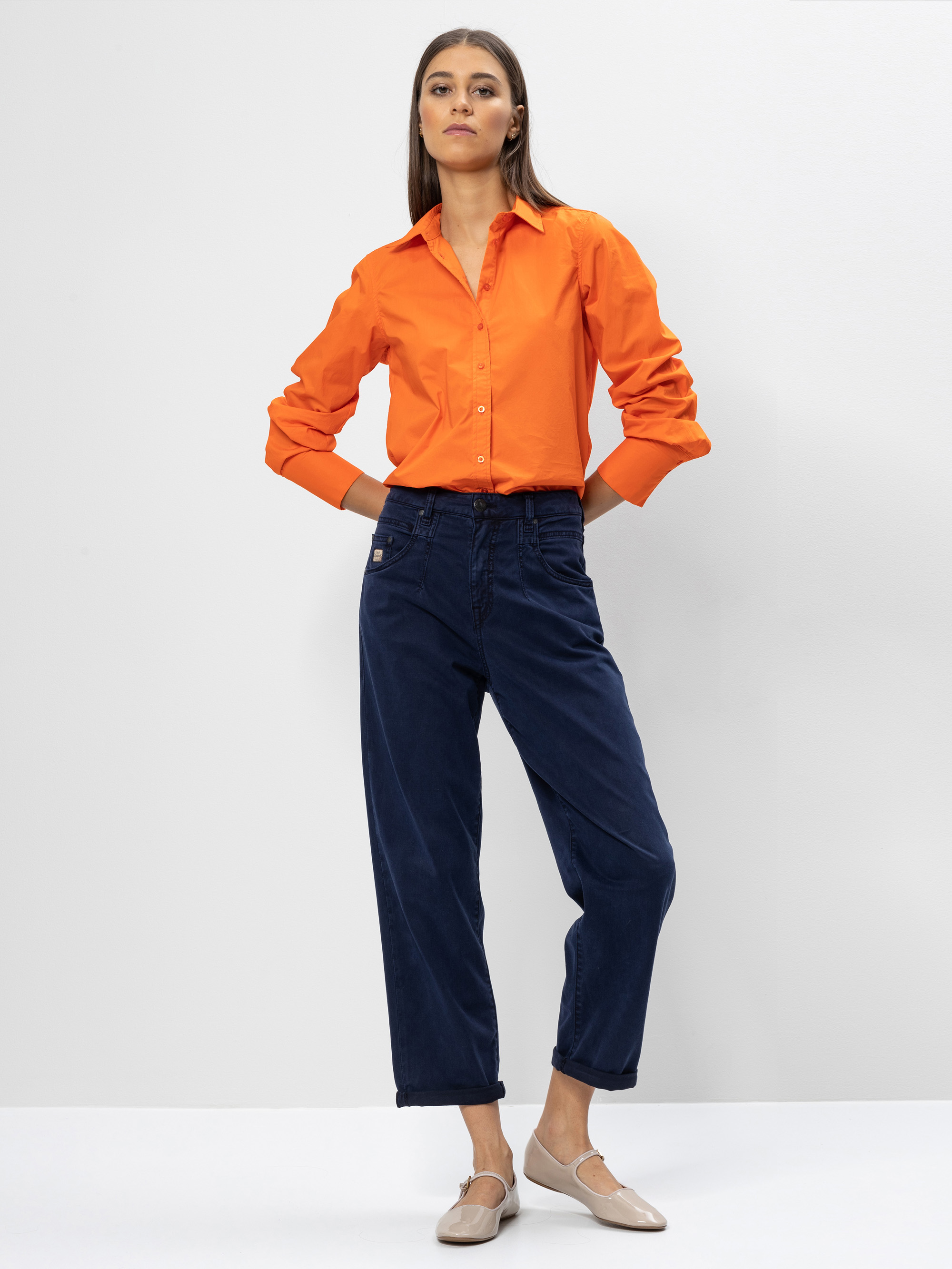 Eine Frau steht vor einem schlichten Hintergrund und trägt ein orangefarbenes Button-up-Hemd, eine dunkelblaue Hose mit hoher Taille und beigefarbene Schuhe. Sie hat glattes braunes Haar und ihre Hände stecken in den Taschen.