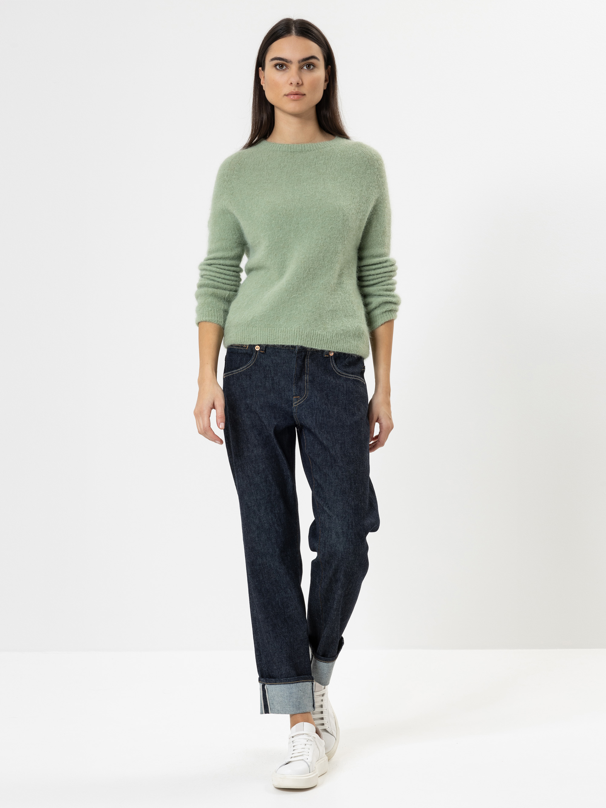 Eine Frau steht vor einem schlichten weißen Hintergrund und trägt einen hellgrünen, wattierten Pullover, dunkle Jeans mit Bündchen und weiße Turnschuhe. Sie hat langes dunkles Haar und einen neutralen Gesichtsausdruck, die Hände stecken in den Tasch