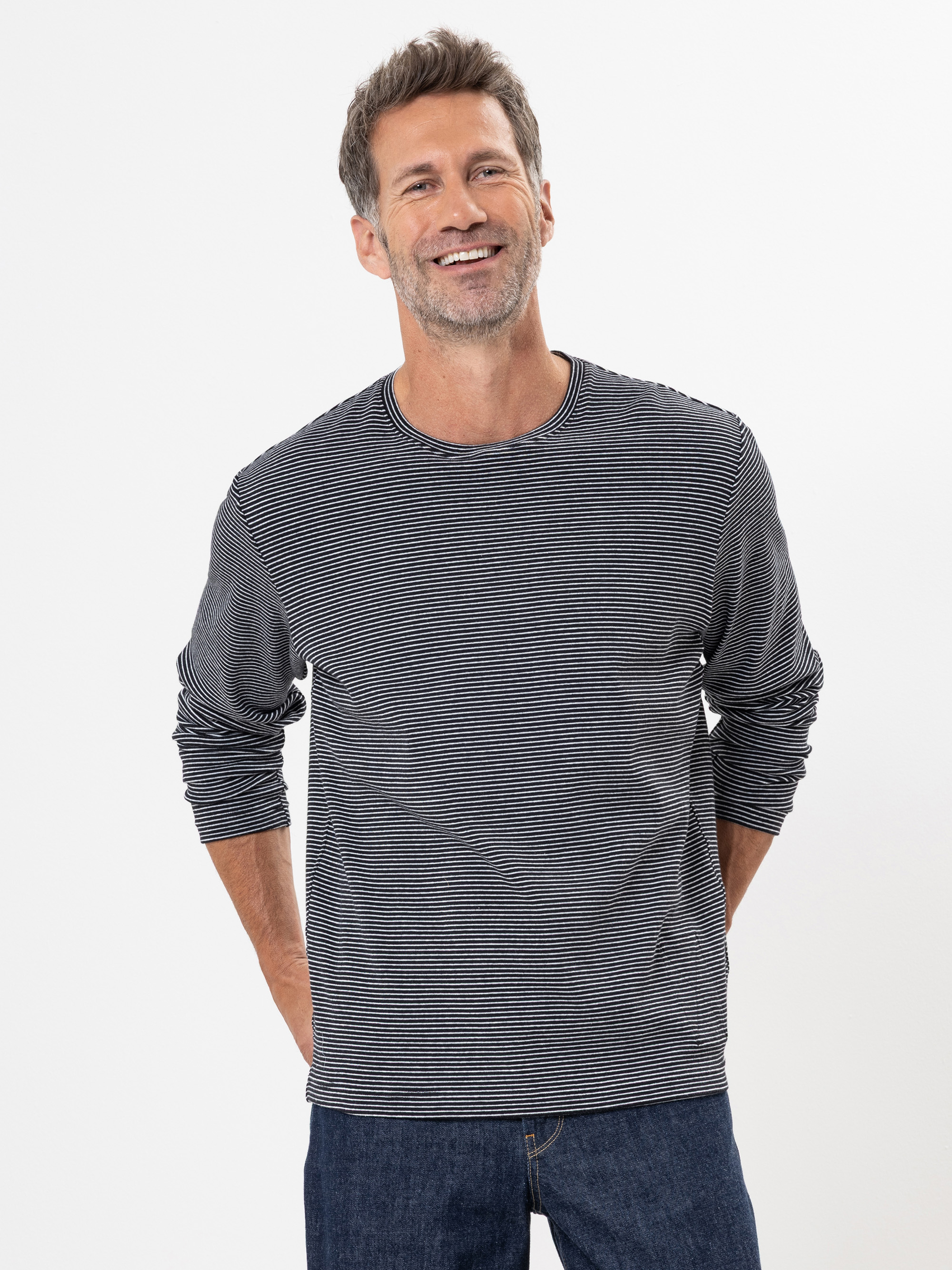 Ein Mann mit kurzen braunen und etwas grauen Haaren steht lächelnd vor einem weißen Hintergrund. Er trägt ein langärmeliges, schwarz-weiß gestreiftes Hemd und eine blaue Jeans, die Hände hinter dem Rücken.