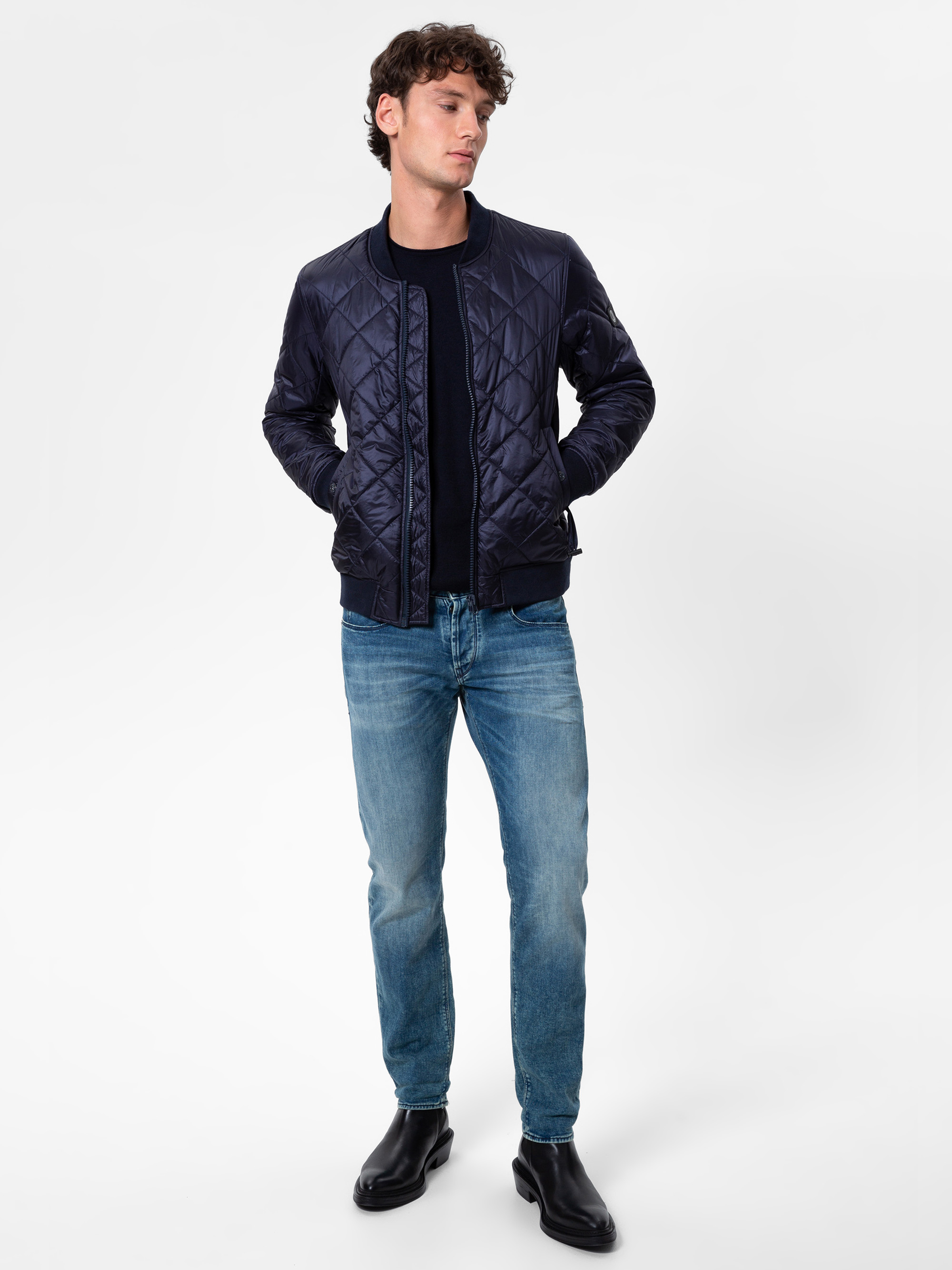 Ein Mann steht vor einem schlichten weißen Hintergrund, trägt eine marineblaue Steppjacke, ein schwarzes Hemd, blaue Jeans und schwarze Stiefel, hat die Hände in den Taschen und schaut leicht zur Seite.