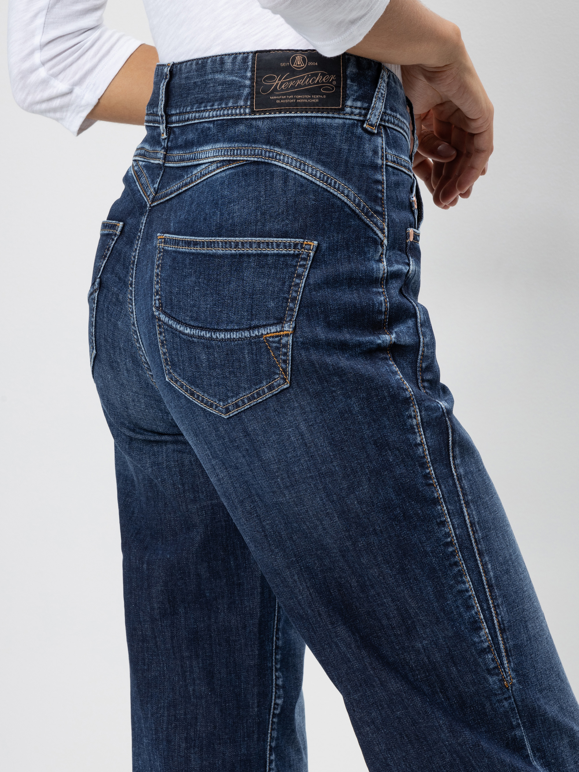 Eine Person, die eine blaue Jeans mit hoher Taille und sichtbaren Nähten und einem Herrlicher-Markenaufnäher am Bund trägt, von der Seite und von hinten gesehen.