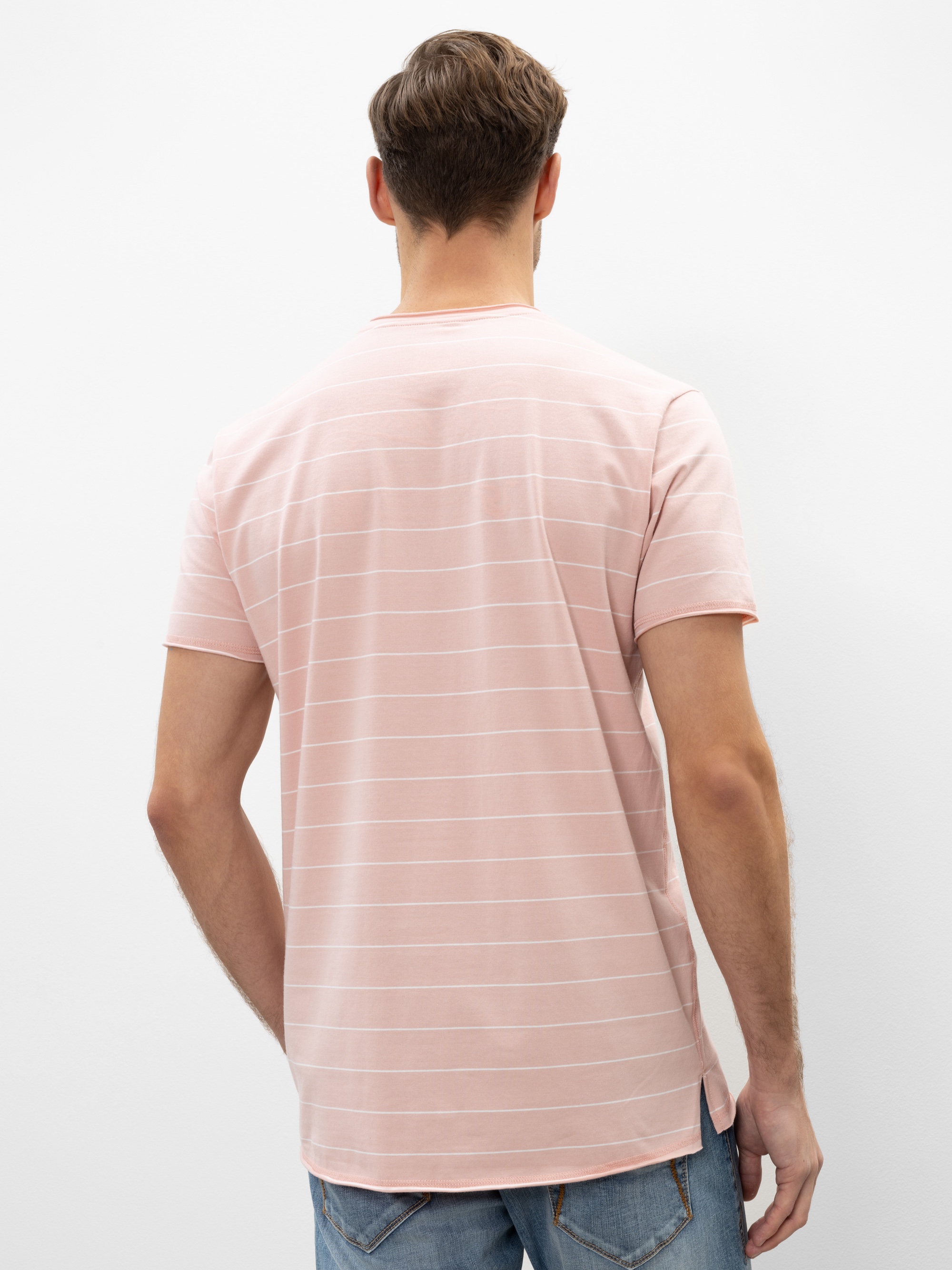 Ein Mann steht mit dem Gesicht zur Wand und trägt ein hellrosa, kurzärmeliges T-Shirt mit dezenten Querstreifen und blaue Jeans. Der Hintergrund ist einfarbig weiß.