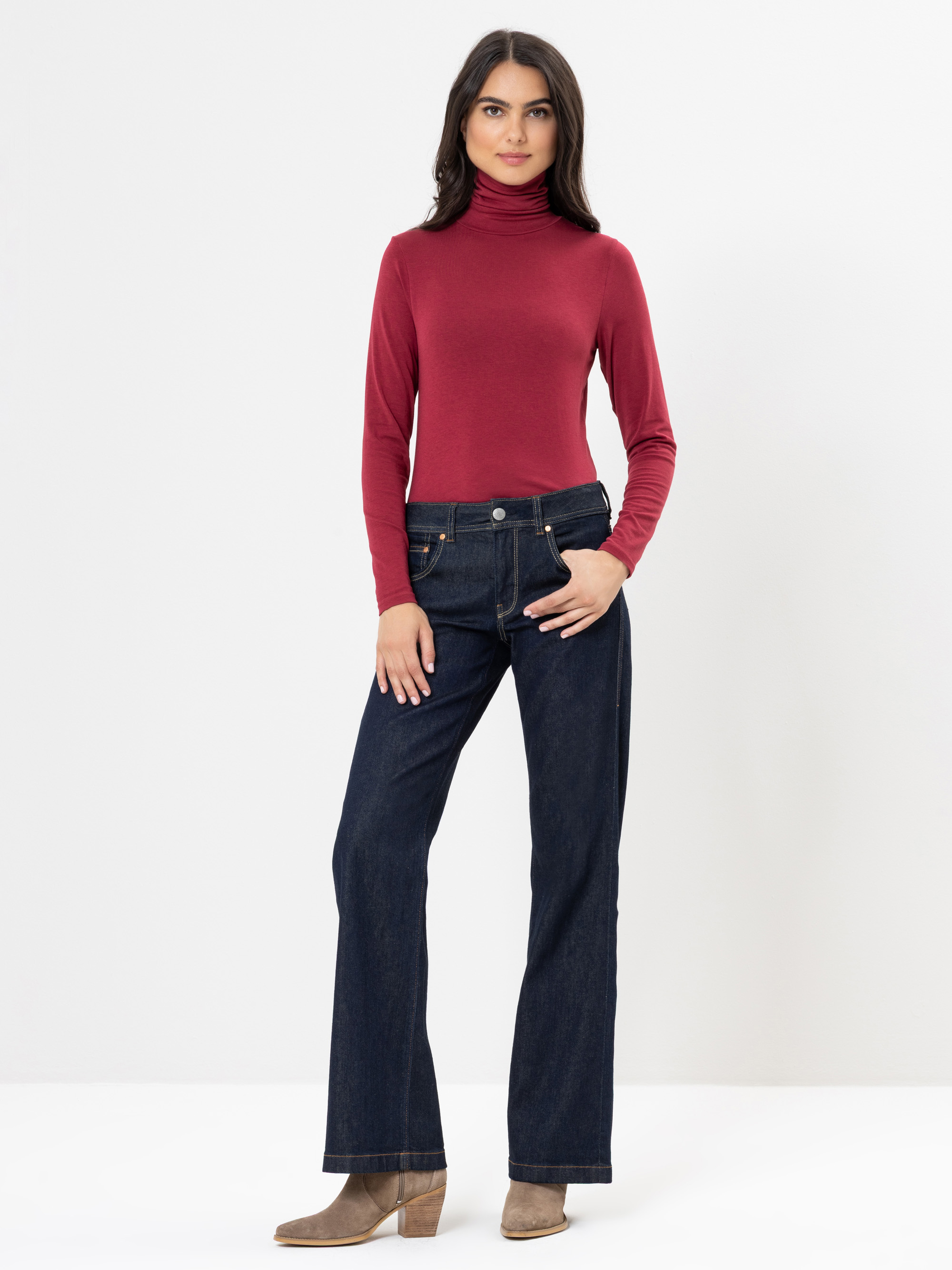 Eine Frau steht vor einem schlichten weißen Hintergrund und trägt einen langärmeligen roten Rollkragenpullover, dunkelblaue Jeans und hellbraune Stiefel mit Absatz. Sie hat langes dunkles Haar und blickt mit einem neutralen Gesichtsausdruck nach vorne.