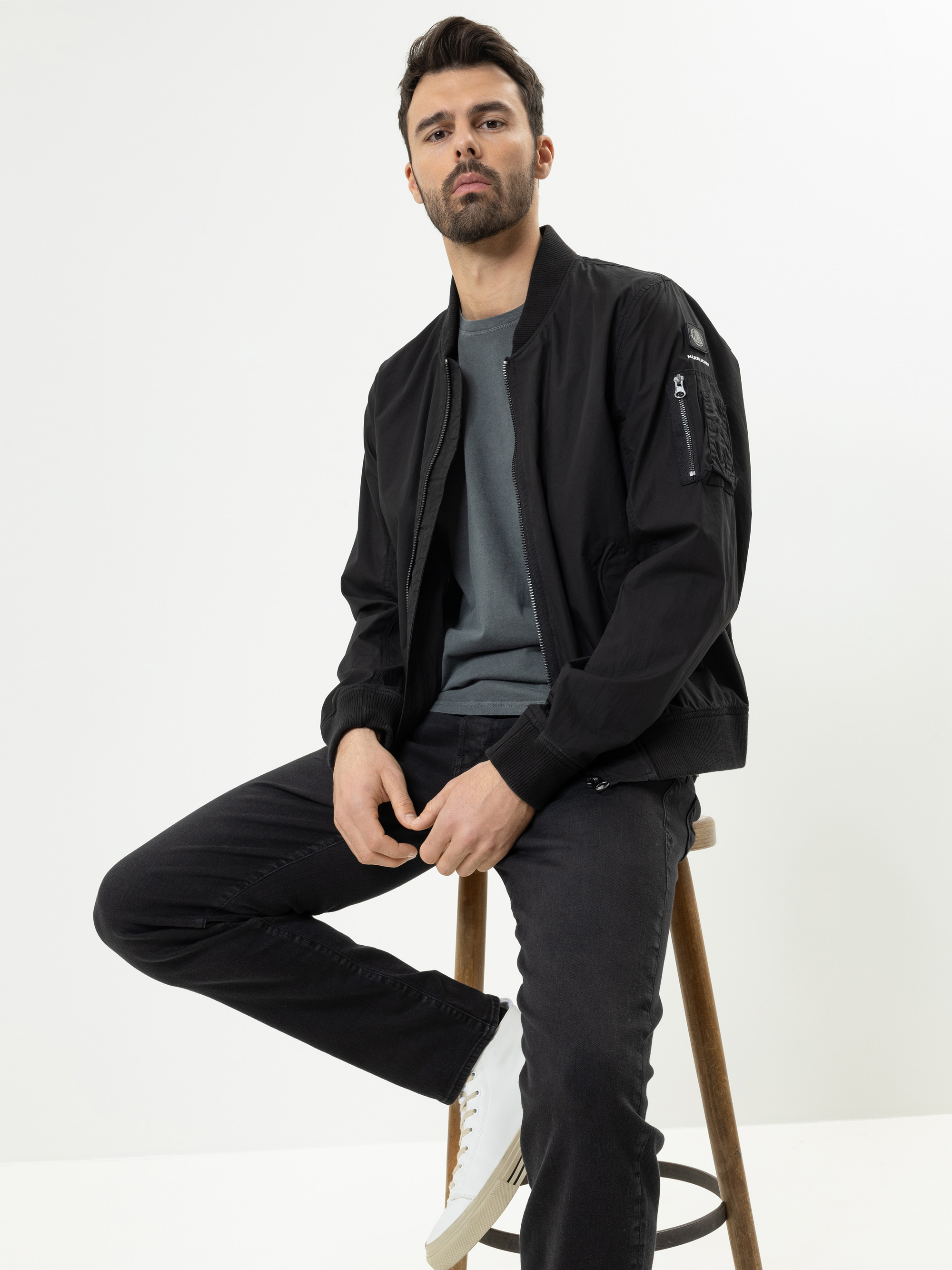 Ein Mann mit kurzen dunklen Haaren und einem Bart sitzt auf einem Holzschemel und trägt eine schwarze Bomberjacke, ein graues Hemd, schwarze Jeans und weiße Turnschuhe vor einem schlichten weißen Hintergrund.