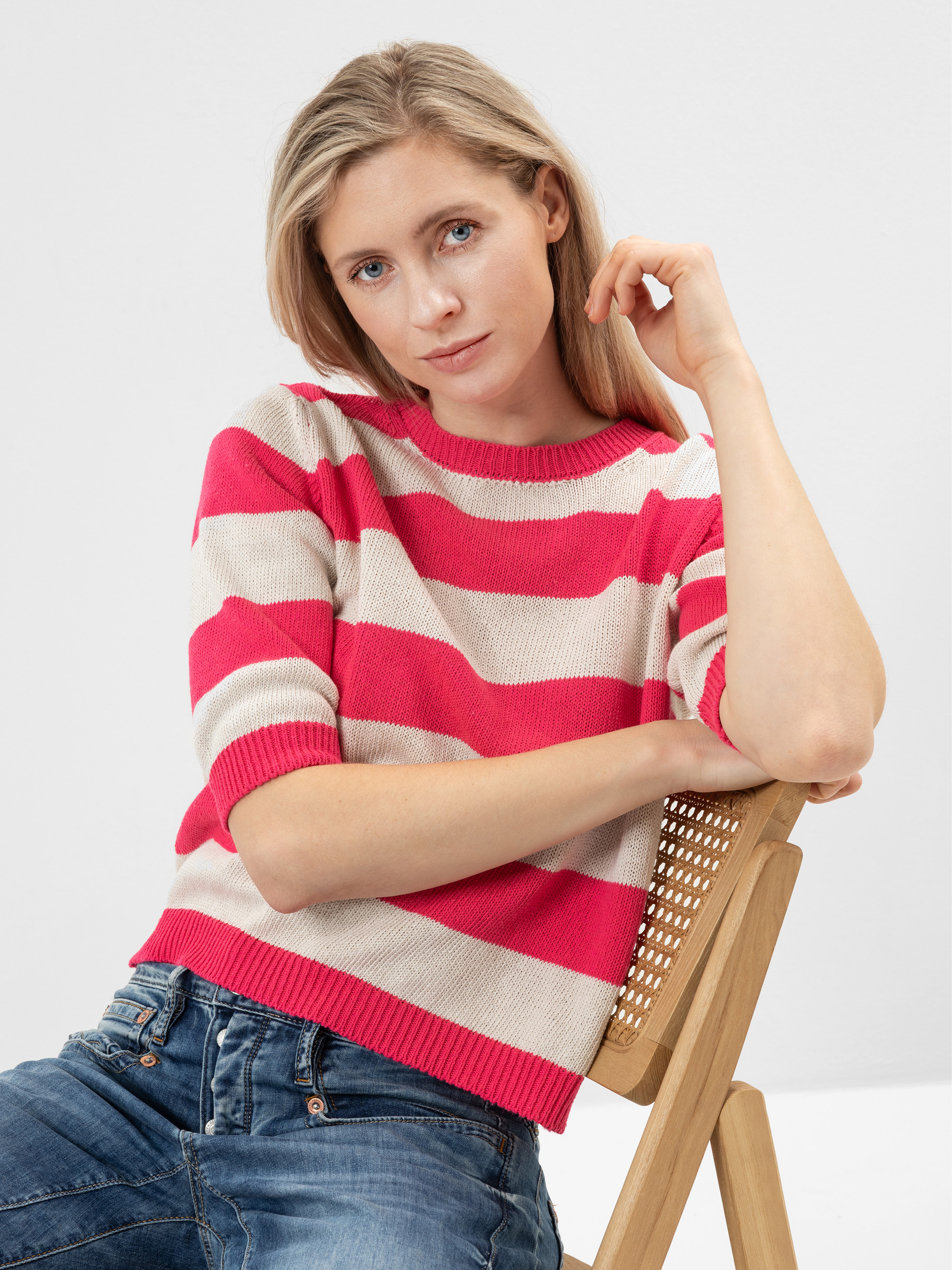 Eine Frau mit blonden Haaren, einem rosa-weiß gestreiften Pullover und blauen Jeans sitzt auf einem Holzstuhl und schaut entspannt in die Kamera.