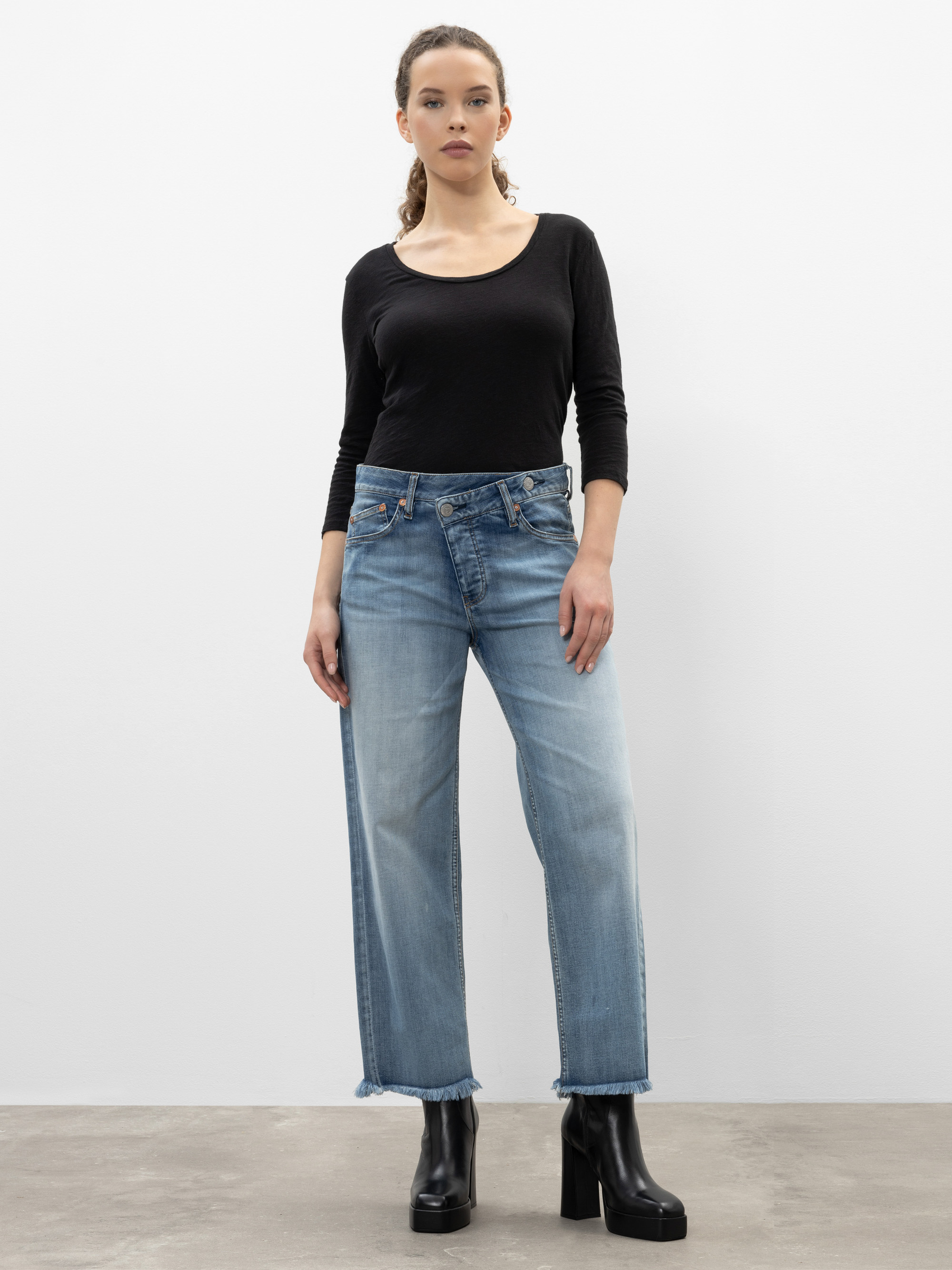 Eine Frau steht vor einer schlichten weißen Wand. Sie trägt ein schwarzes, langärmeliges Oberteil, hellblaue Jeans mit geradem Bein und schwarze, hochhackige Stiefel. Sie blickt nach vorne und hat die Arme entspannt an den Seiten.