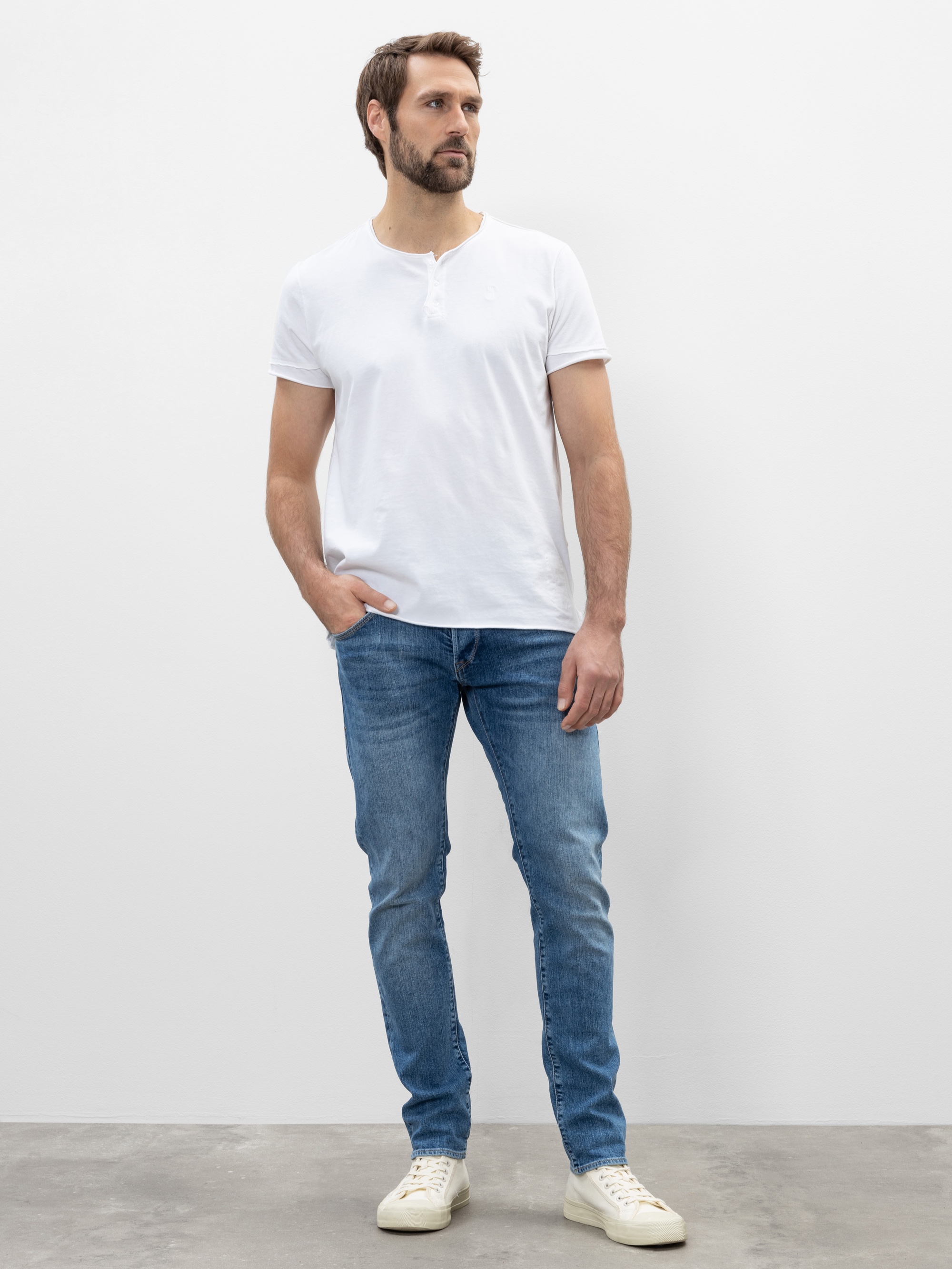 Ein Mann mit kurzen braunen Haaren und Bart steht vor einer schlichten weißen Wand. Er trägt ein weißes, kurzärmeliges Henley-Shirt, blaue Jeans und weiße Turnschuhe und hat eine Hand in der Tasche.