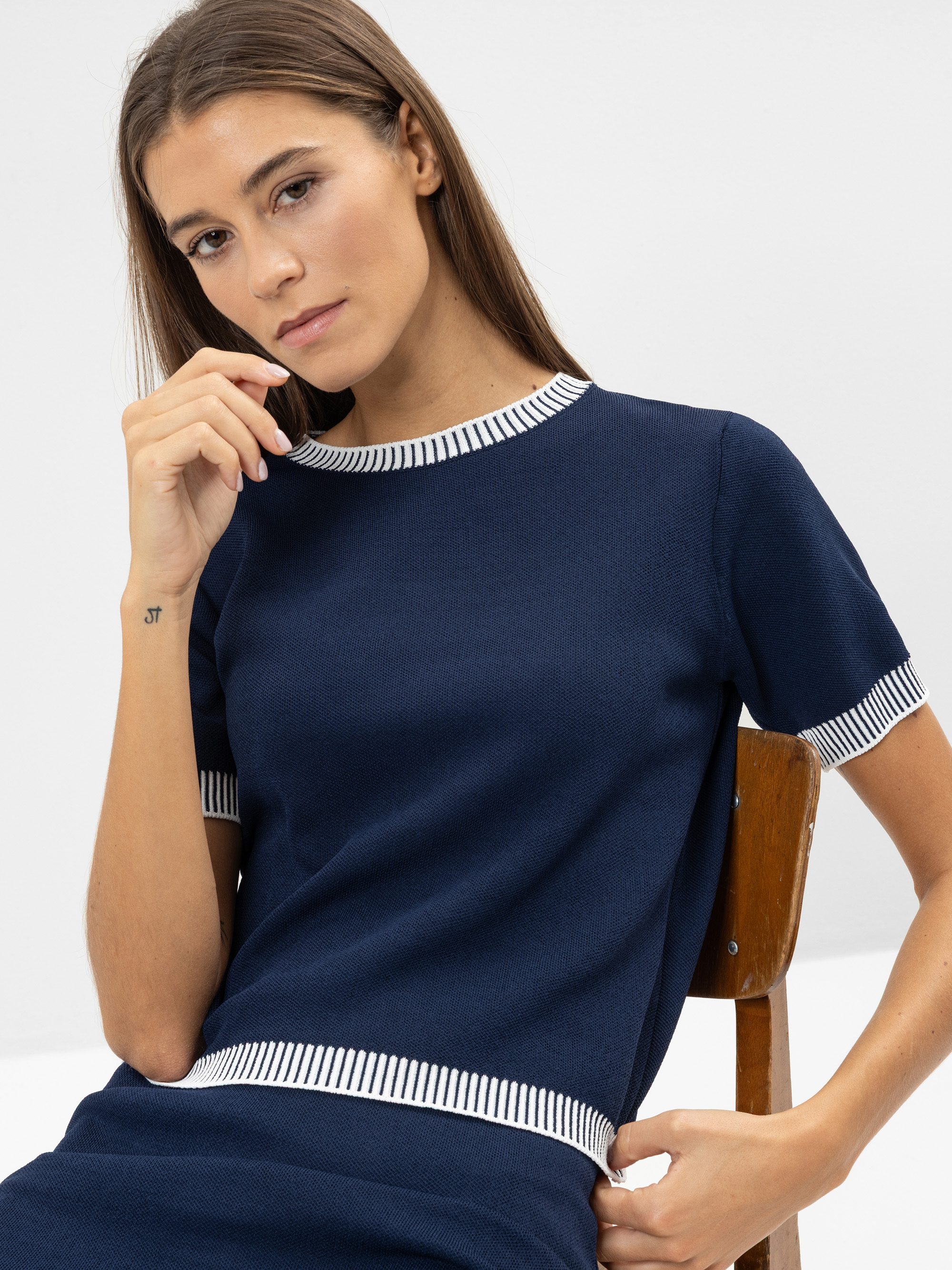 Eine Frau mit langen braunen Haaren sitzt auf einem Holzstuhl und trägt ein marineblaues, kurzärmeliges Oberteil mit weiß-marinefarbenen Streifen an Kragen und Manschetten. Sie hält ihre linke Hand neben ihr Gesicht und hat eine kleine Tätowierung au